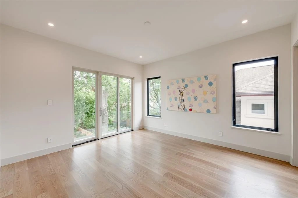 Empty living room with hardwood floors, white walls, large glass sliding door, and two windows, with a colorful art piece on the wall.