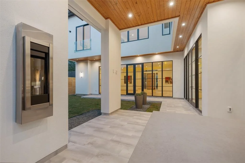 Modern house patio with white walls, wooden ceiling, glass doors, and potted plants, illuminated by recessed lighting.