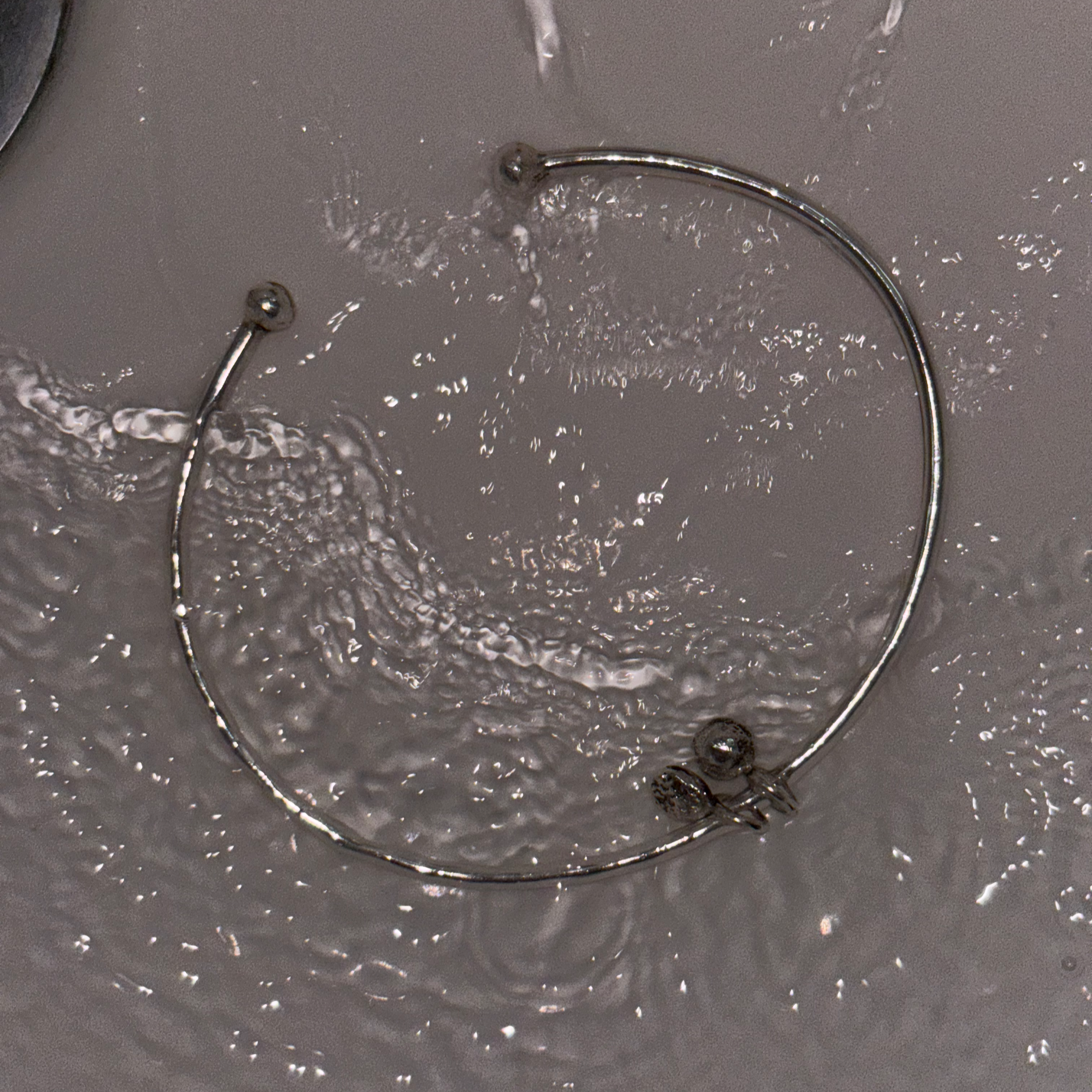 The Charm Open Bangle is submbered in running water over a white background. The flash of the camera over the polish of the sterling silver causes ripples of light to reflect in the water.