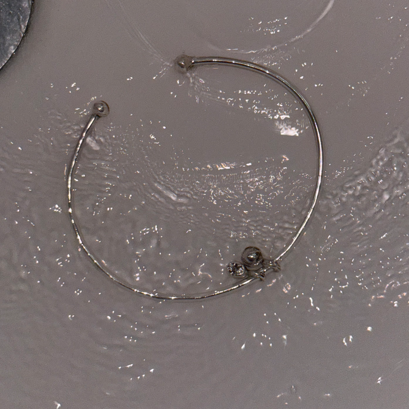 The Charm Open Bangle is submbered in running water over a white background. The flash of the camera over the polish of the sterling silver causes ripples of light to reflect in the water.