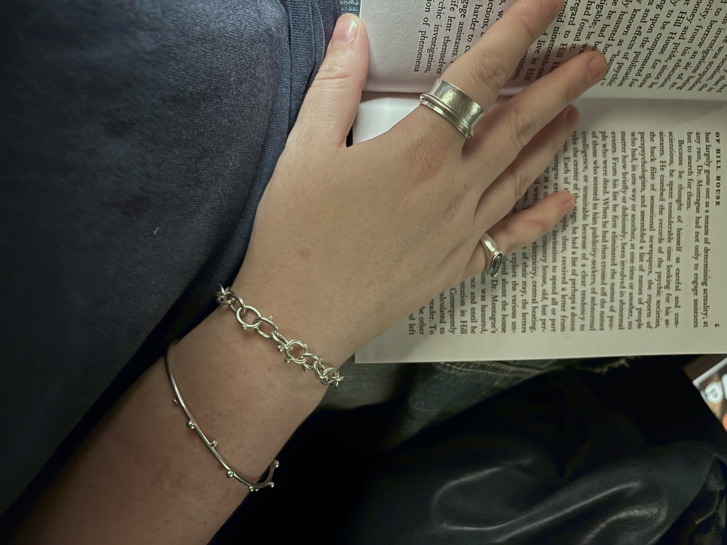 A hand adorned with The Spinner Ring, The Blobby Bangle and the Blobby Chain, lies on top of an open book.