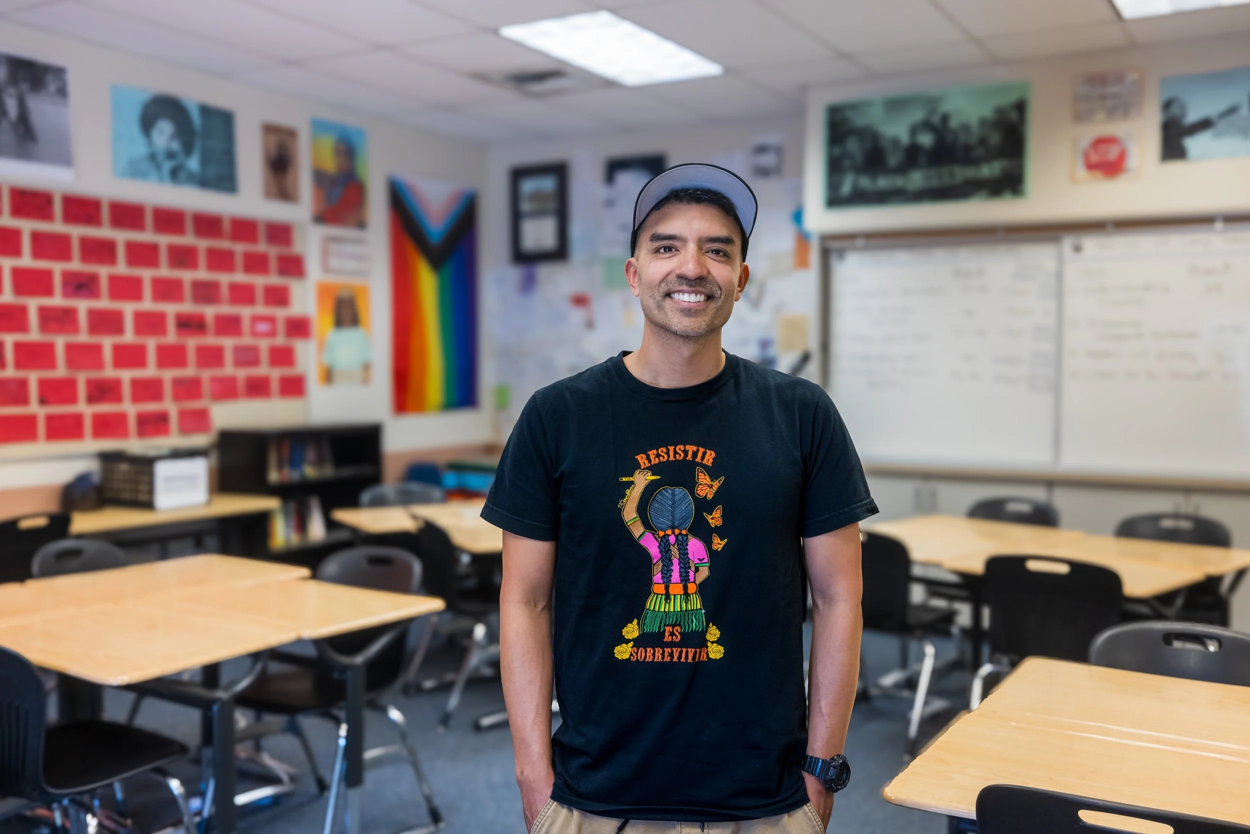 A smiling man wearing a black T-shirt and a cap standing in a classroom with tables, chairs, and colorful posters on the walls.