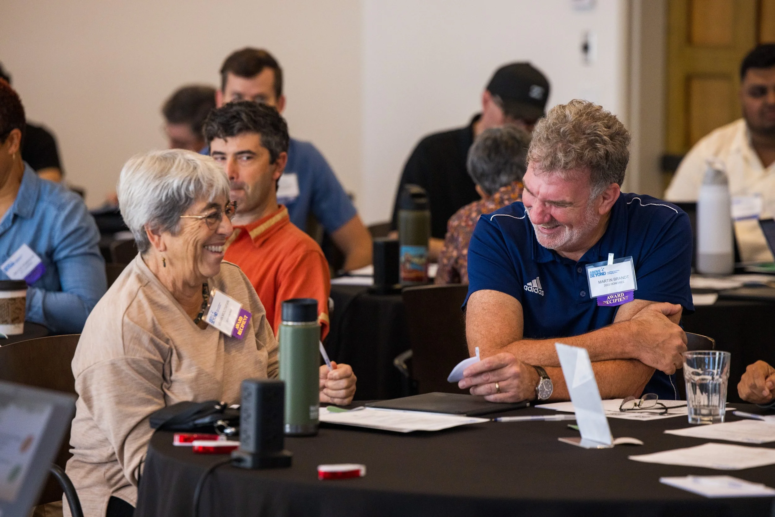 Two people, an elderly woman and a middle-aged man, smiling and engaging in conversation at a conference table with papers, water bottles, and name tags.