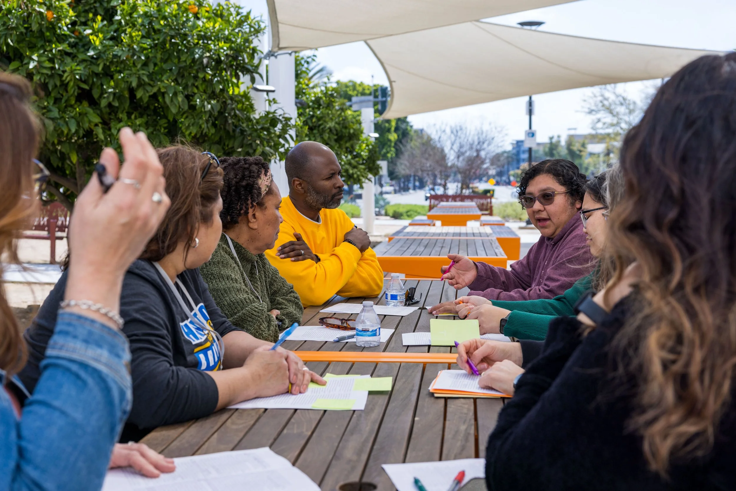 A group of diverse people sitting around a long outdoor table, engaged in a discussion during daytime, under a large shade structure with trees and street visible in the background.