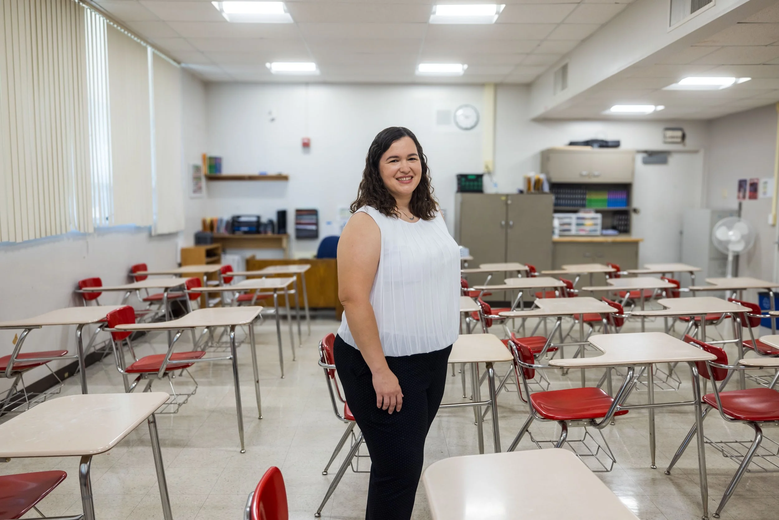 A woman standing in a classroom with red desks and chairs, smiling at the camera.