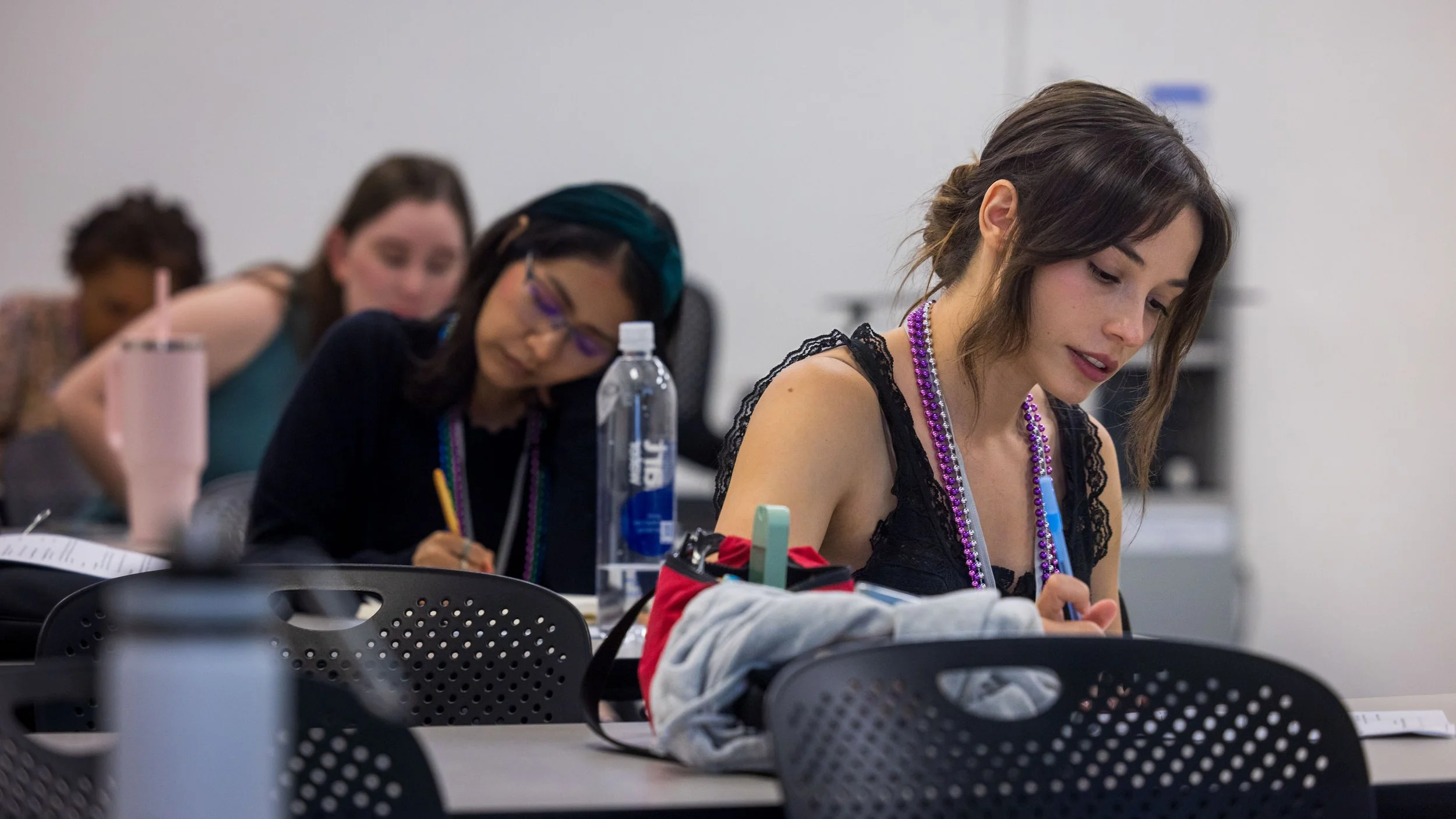 Multiple women sitting at desks focusing on their work in a classroom or workshop setting, with various items like water bottles and notebooks around.