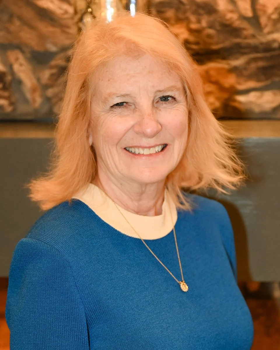 A smiling woman with light skin, red hair, and blue eyes wearing a blue top with a cream collar and a gold necklace, sitting indoors against a brown, textured background.