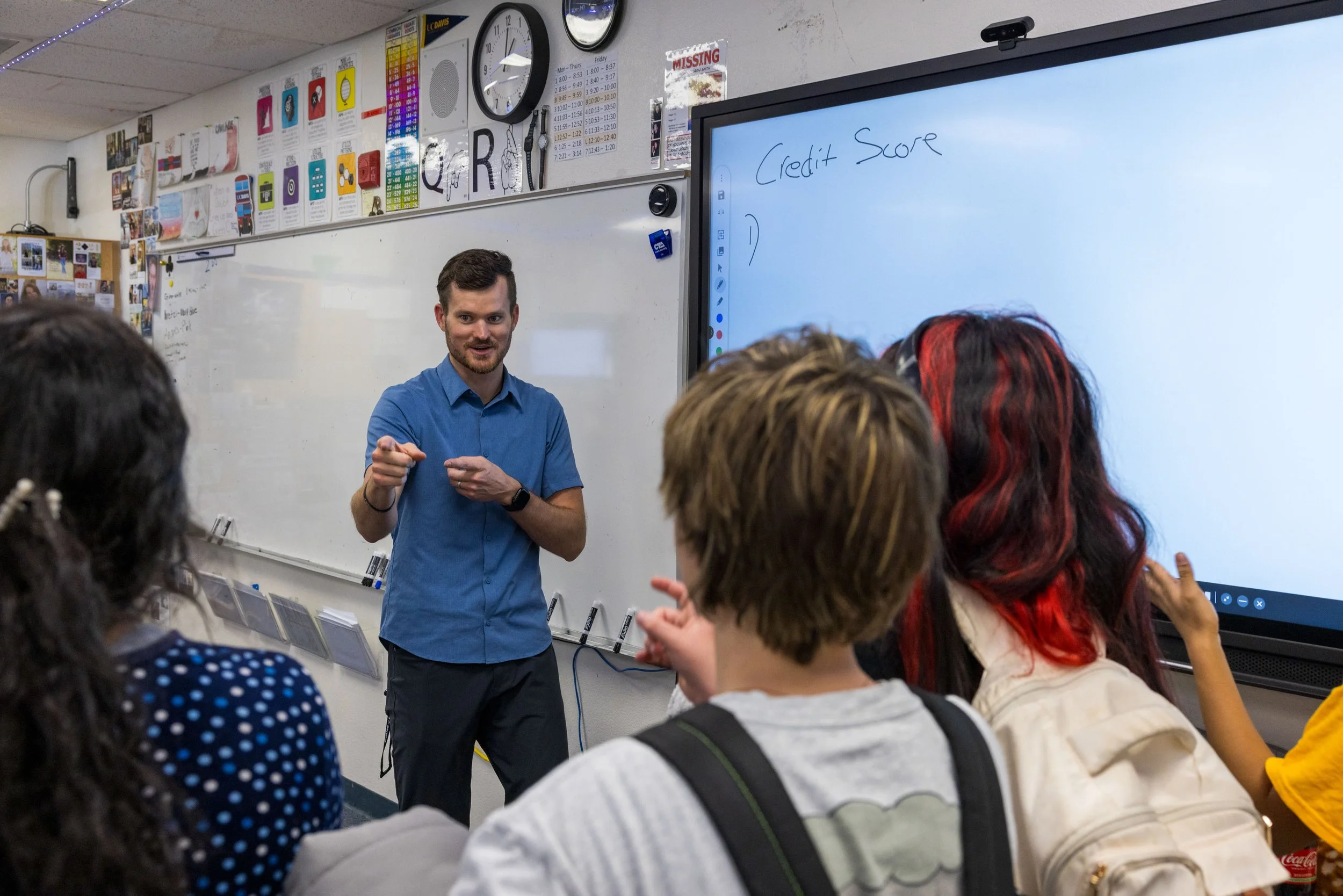 A teacher standing in front of a classroom teaches a group of students. The teacher is gesturing with his hands, and there is a digital whiteboard with the words 'Credit Score' written on it. Students are attentively listening, with some raising their hands.