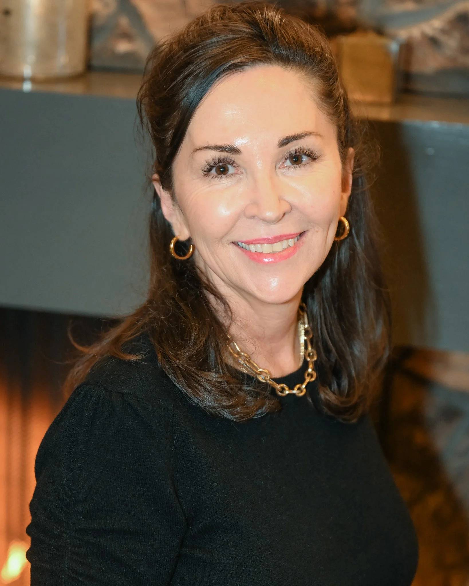 A woman with shoulder-length brown hair, wearing gold hoop earrings, a gold chain necklace, and a black top, smiling at the camera.
