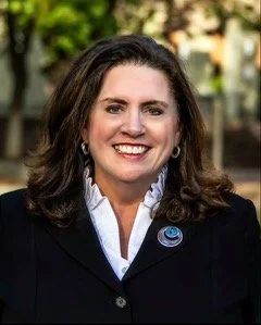 Portrait of a woman with shoulder-length brown hair, wearing a dark blazer and white shirt, smiling outdoors with trees in the background.