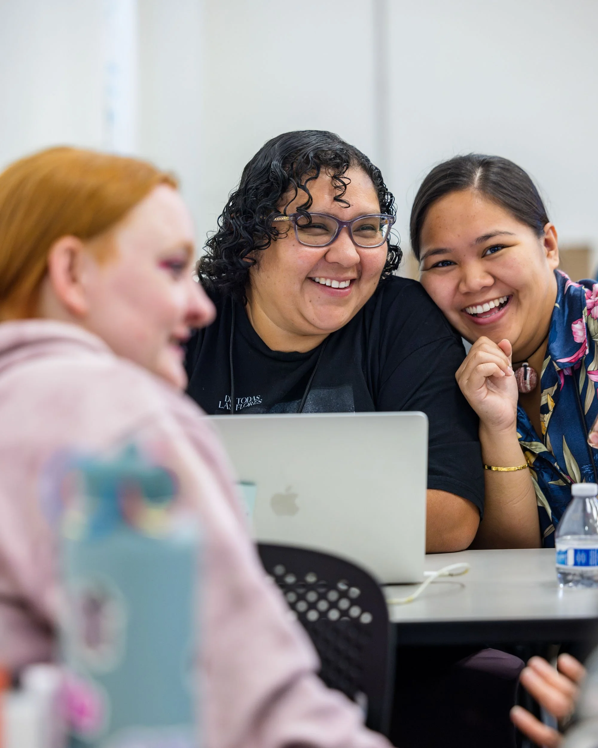 Three women laughing and smiling together at a table with a laptop and a water bottle.