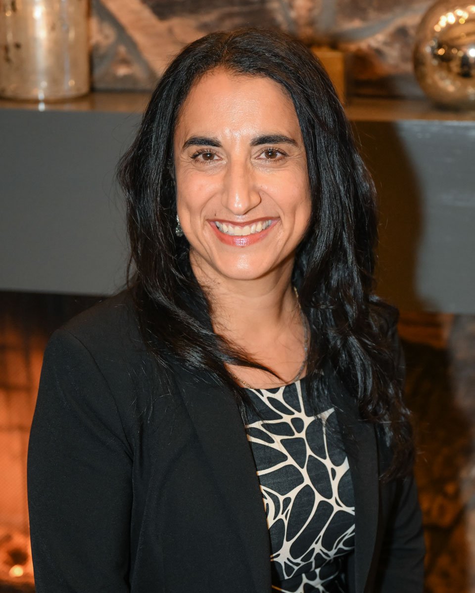 A woman with long dark hair, smiling, wearing a black blazer over a patterned top, in an indoor setting with warm lighting and decorative objects in the background.