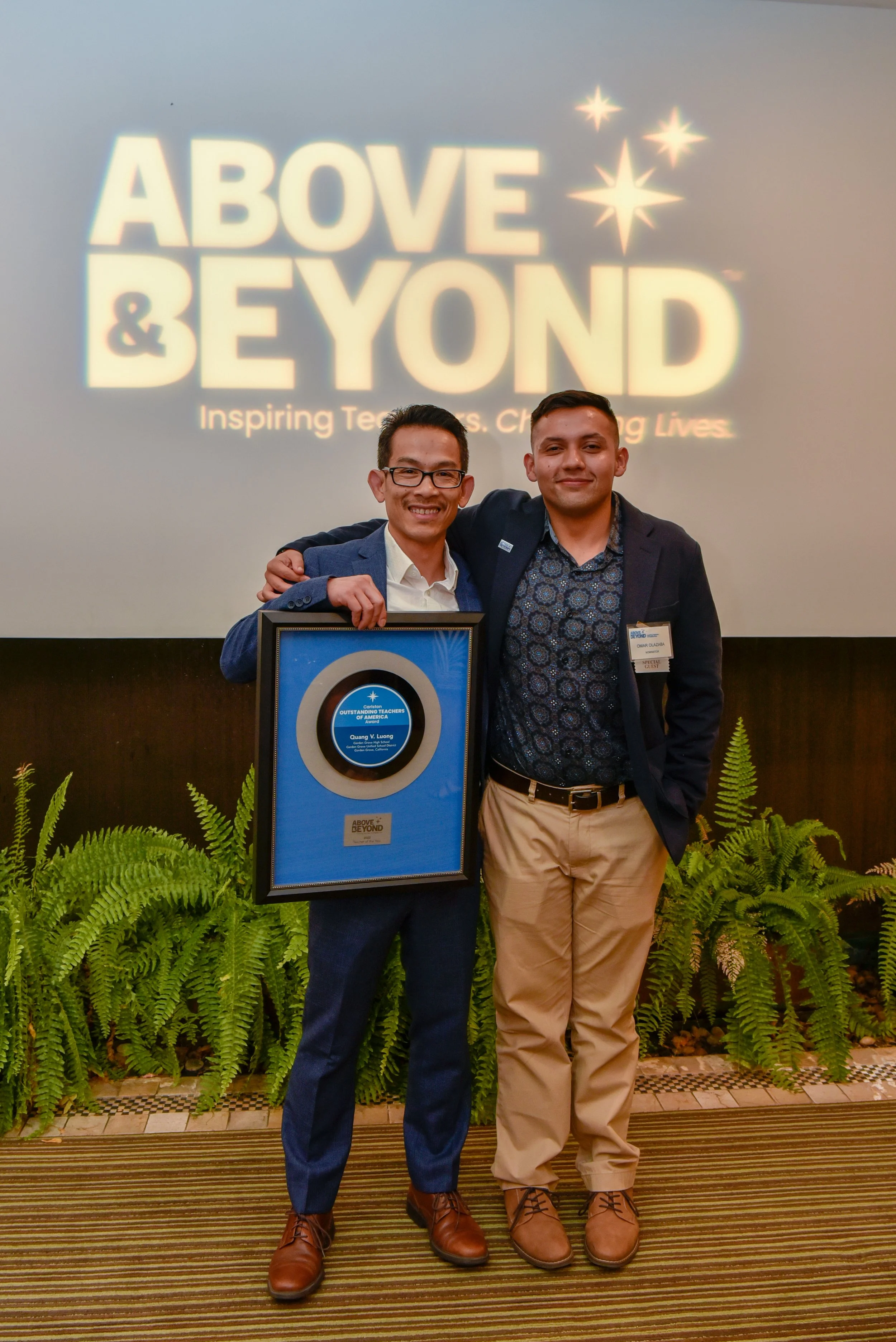 Two men standing on a stage at an award ceremony, one holding a framed award, with a large illuminated "Above & Beyond" sign in the background. The men are smiling and have their arms around each other, surrounded by green plants.