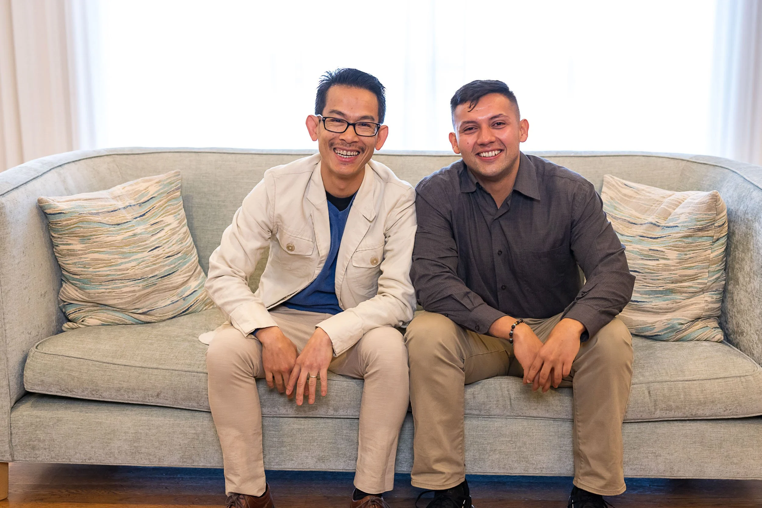 Two men sitting on a beige sofa with patterned pillows, smiling at the camera in a well-lit room.