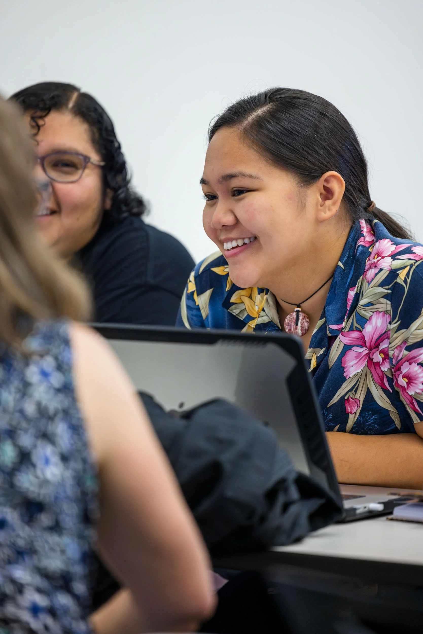 Three women are sitting at a table smiling and engaging in conversation, with one woman in focus wearing a colorful floral shirt and a necklace.