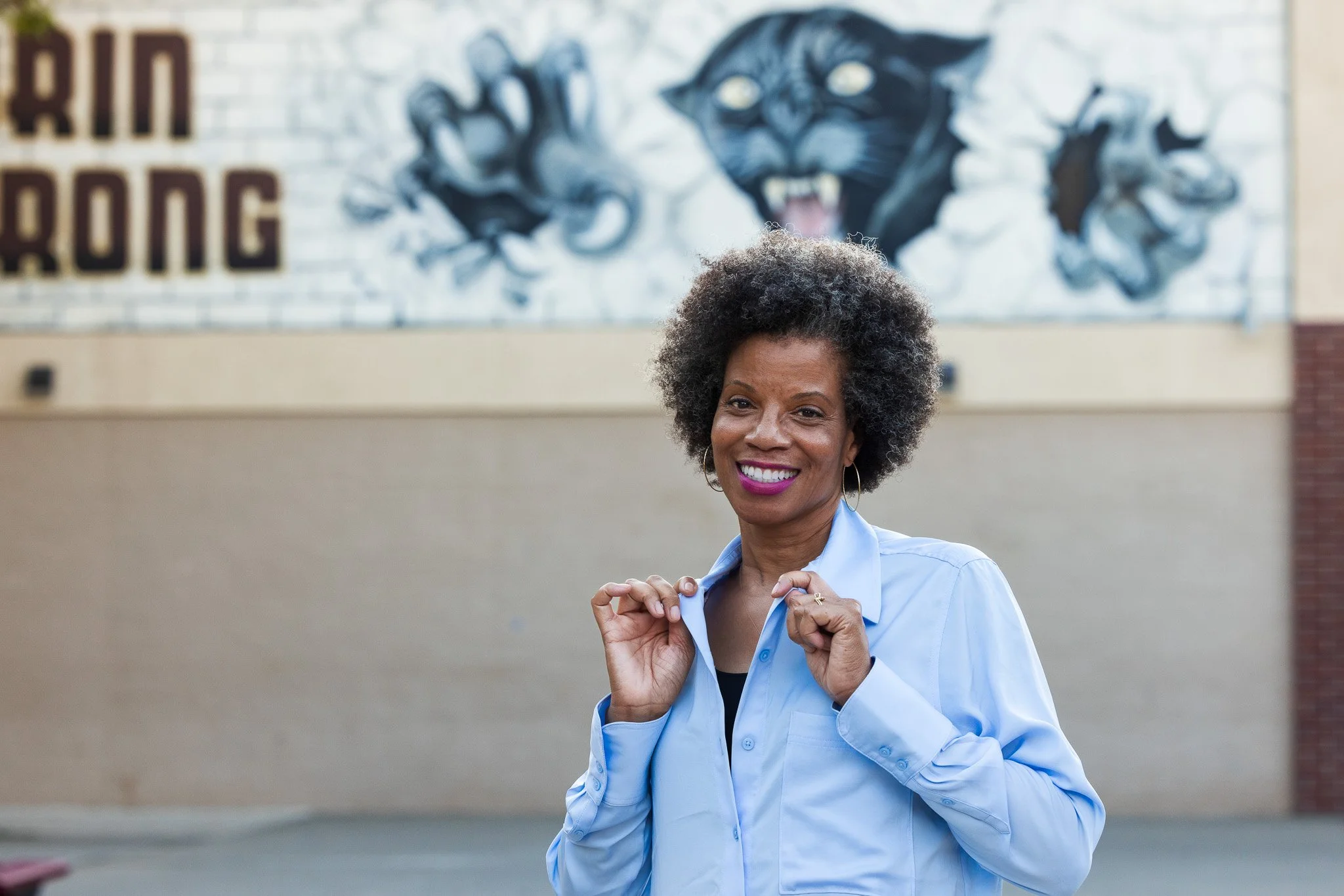 A woman smiling and holding the collar of her light blue shirt in front of a mural featuring a black panther with its paws reaching forward and the partial text 'RIN' and 'BONG' in the background.