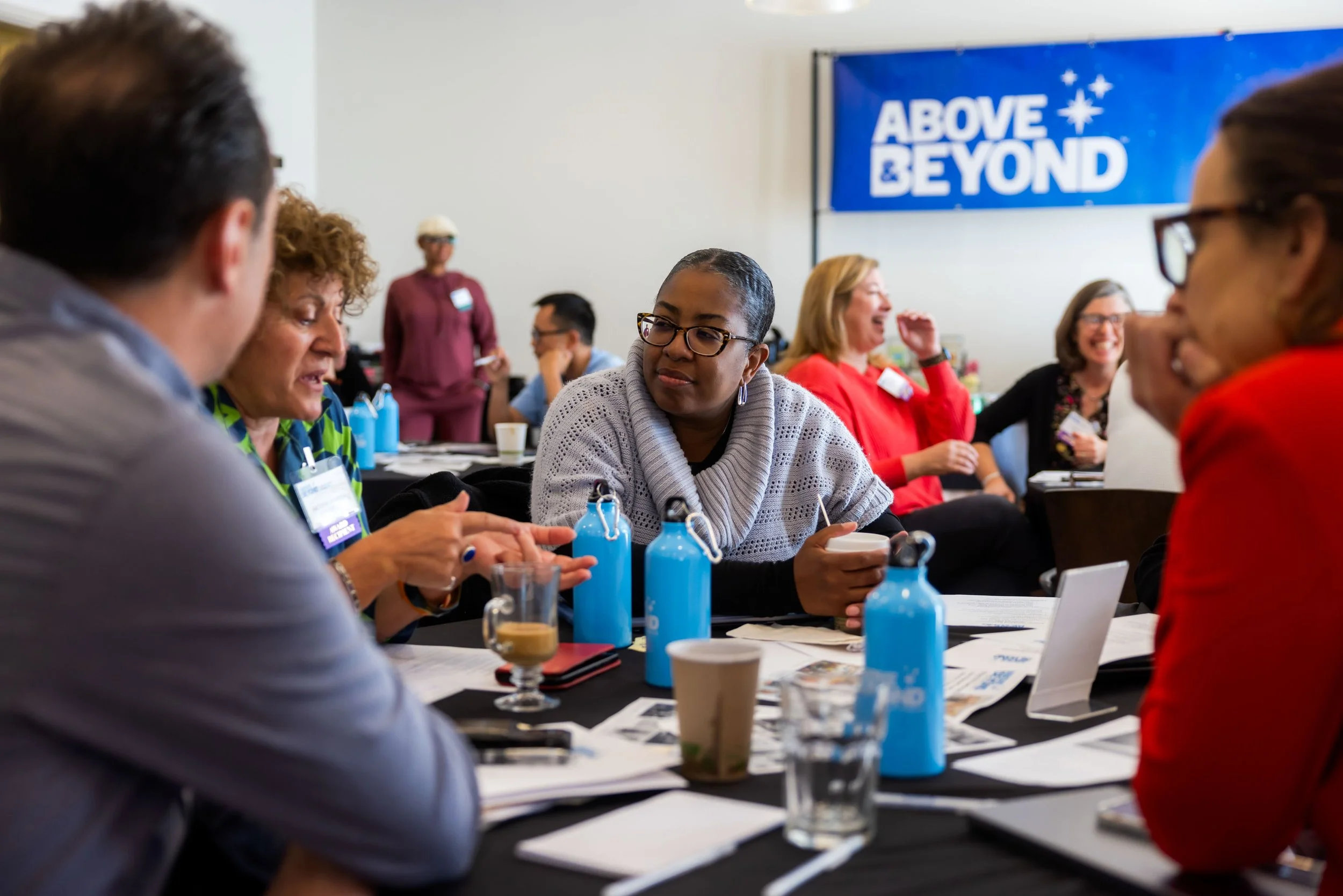 Group of women sitting at a conference table engaged in conversation, with some smiling and others listening. A blue banner with white text 'ABOVE & BEYOND' is visible in the background.