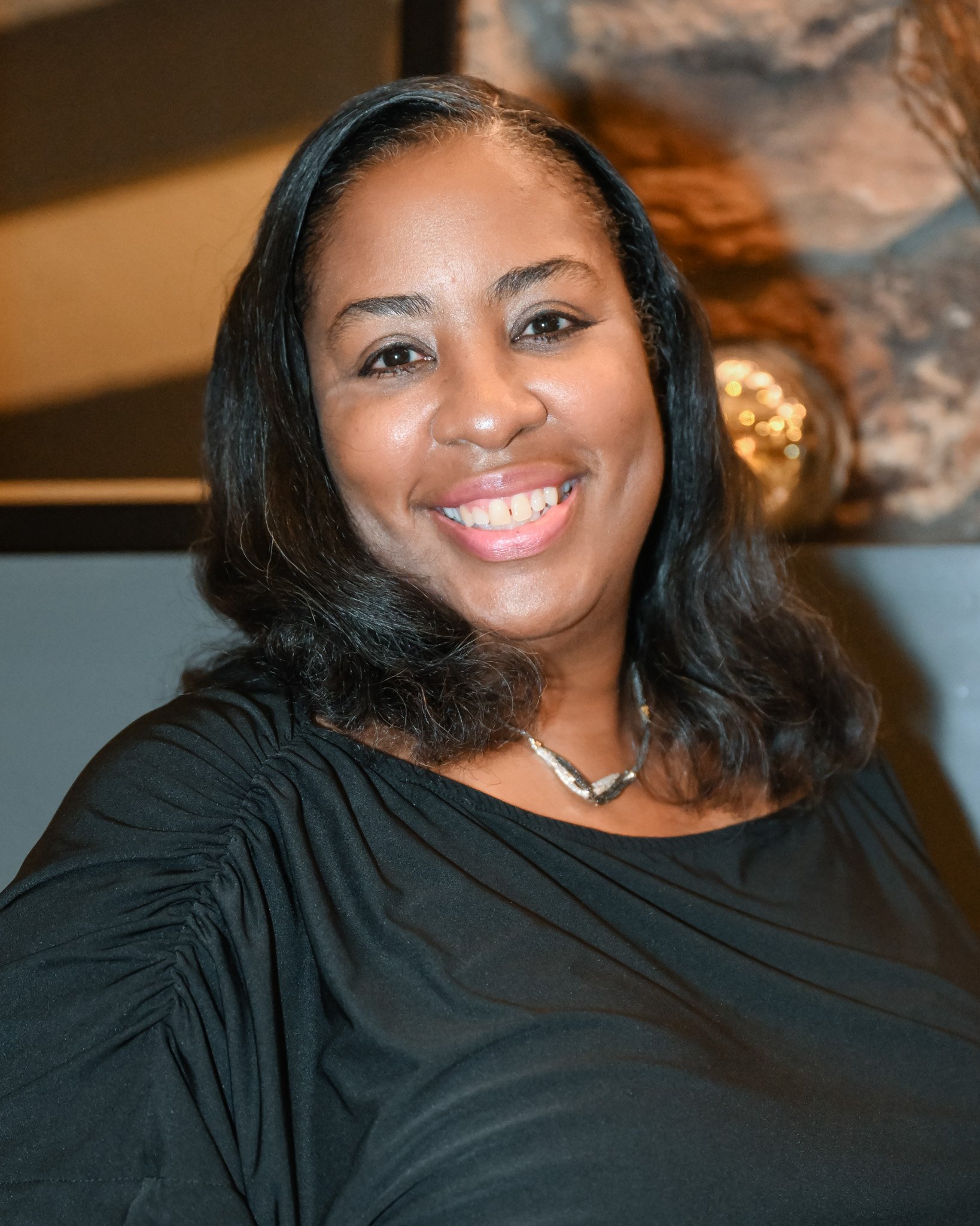 A smiling woman with shoulder-length black hair, wearing a black dress and a necklace, in front of a background with warm lighting and decorative elements.
