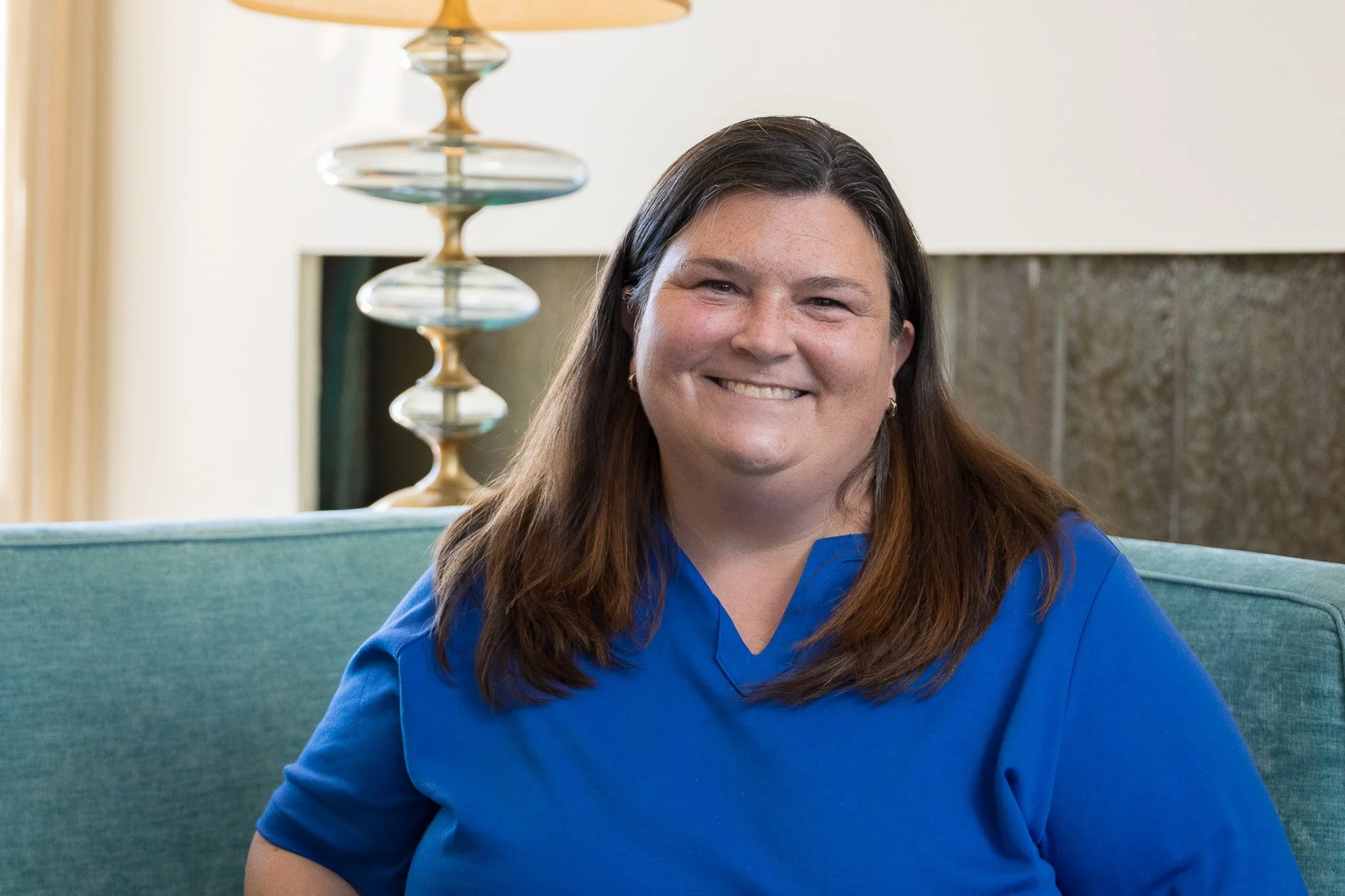 Smiling woman with long brown hair wearing a blue shirt sitting on a teal couch in a well-lit room.