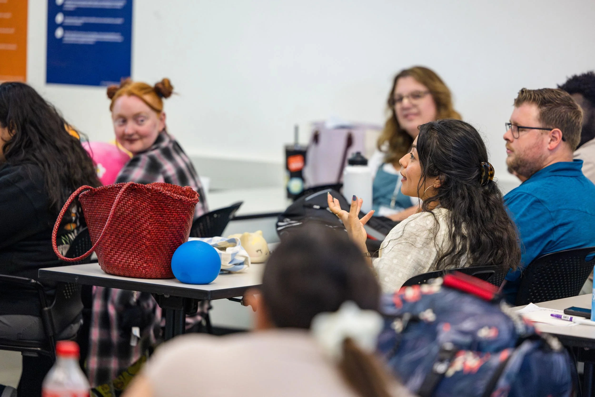 People sitting at desks in a classroom or meeting, engaging in conversation, with bags and personal items on the tables.
