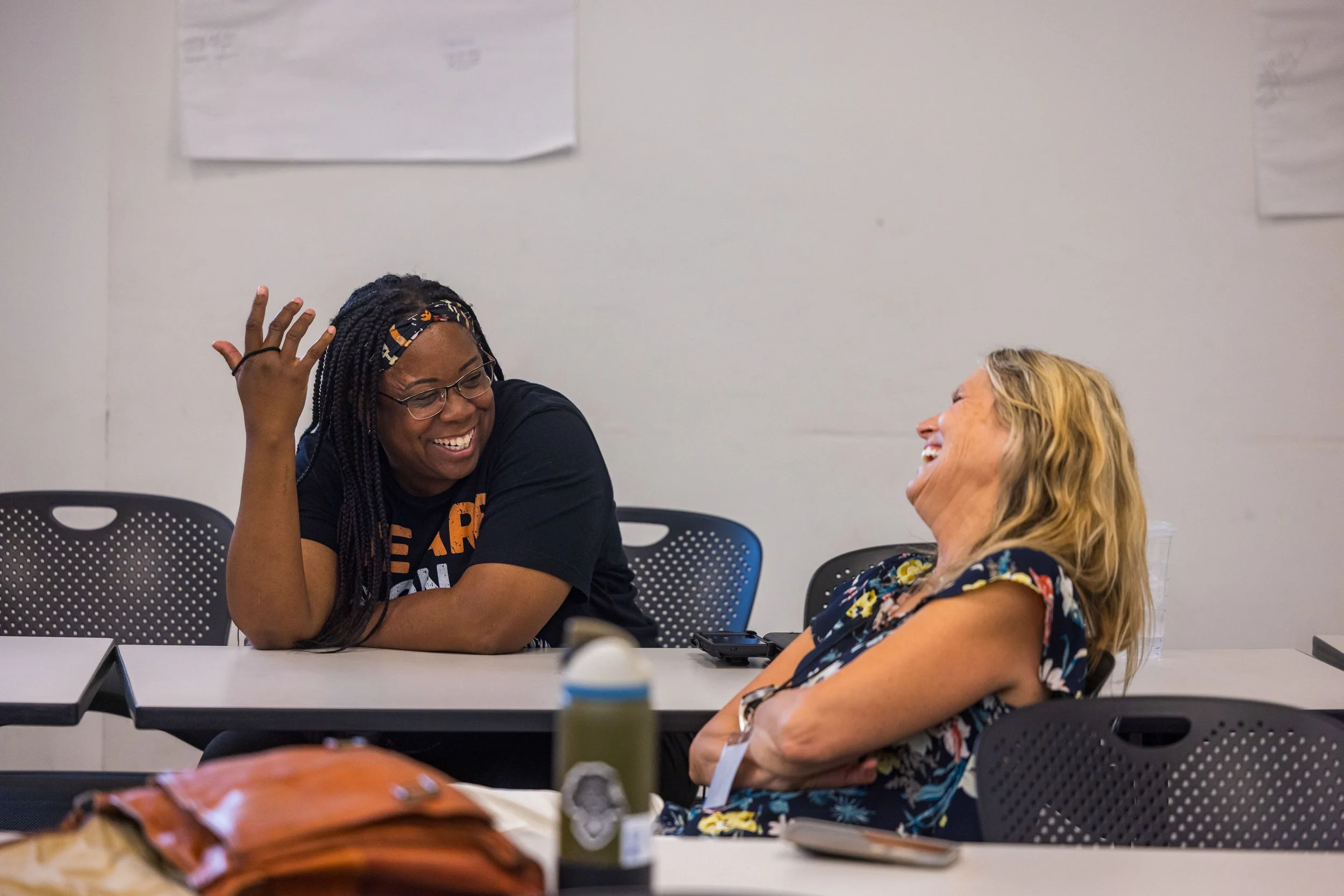Two women are sitting at a table and laughing together, one woman has dark skin, wearing glasses and a black shirt, and the other has light skin, blonde hair, and a floral dress.