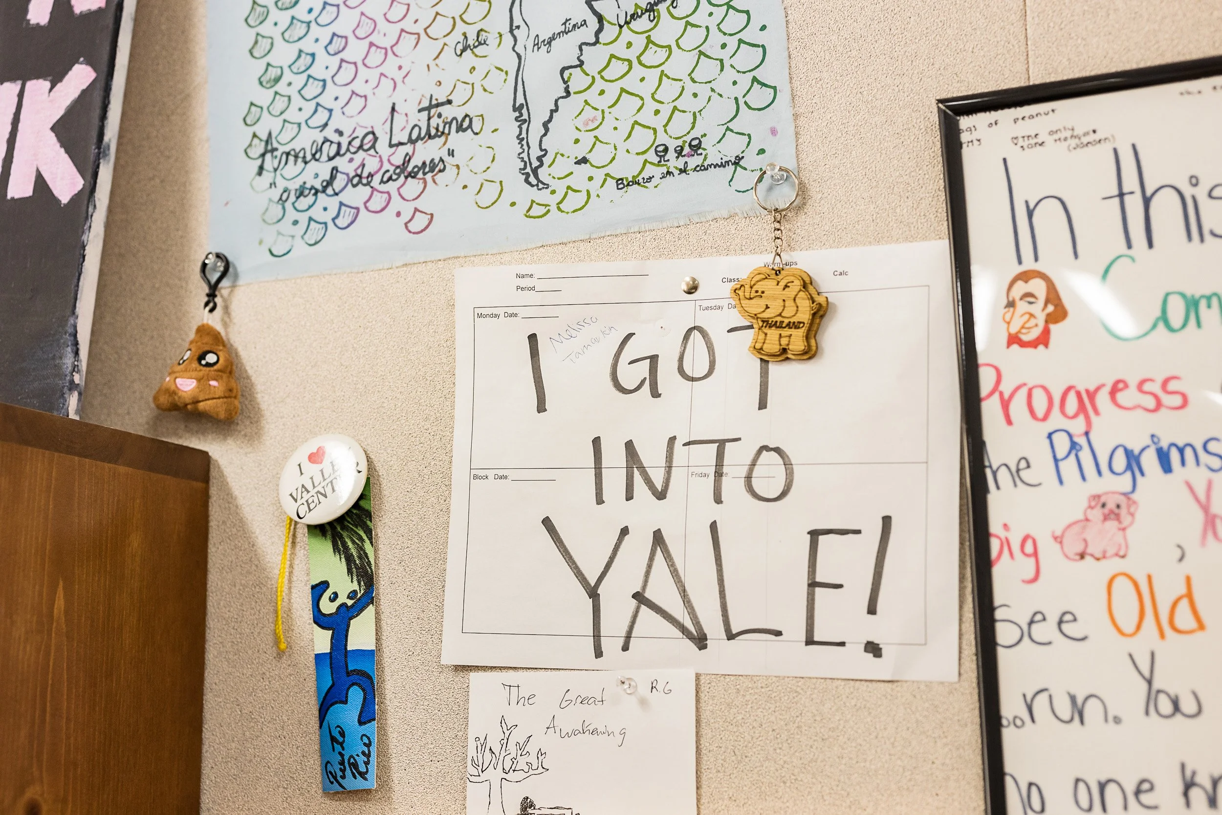 A wall bulletin board with various decorations and writings, including a large hand-drawn map of South America with countries labeled, a plush poop emoji and a Thai elephant keychain hanging from thumbtacks, a button with heart and text, a bookmark w
