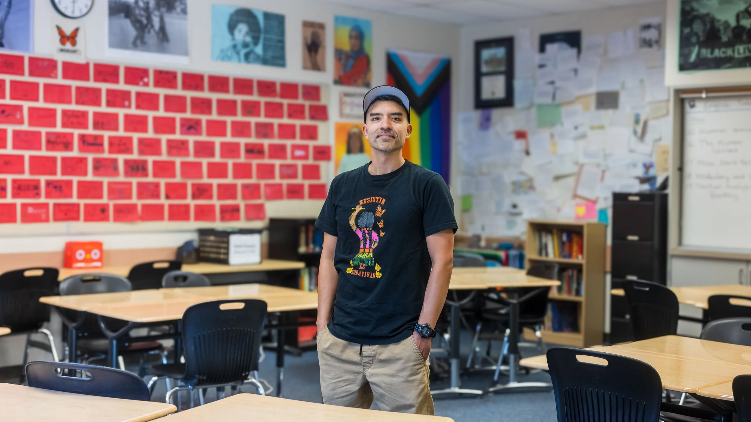 A young man with dark hair and a beard standing in a classroom with colorful posters and papers on the walls. He is wearing a black t-shirt with a graphic and tan cargo pants, with hands in his pockets.