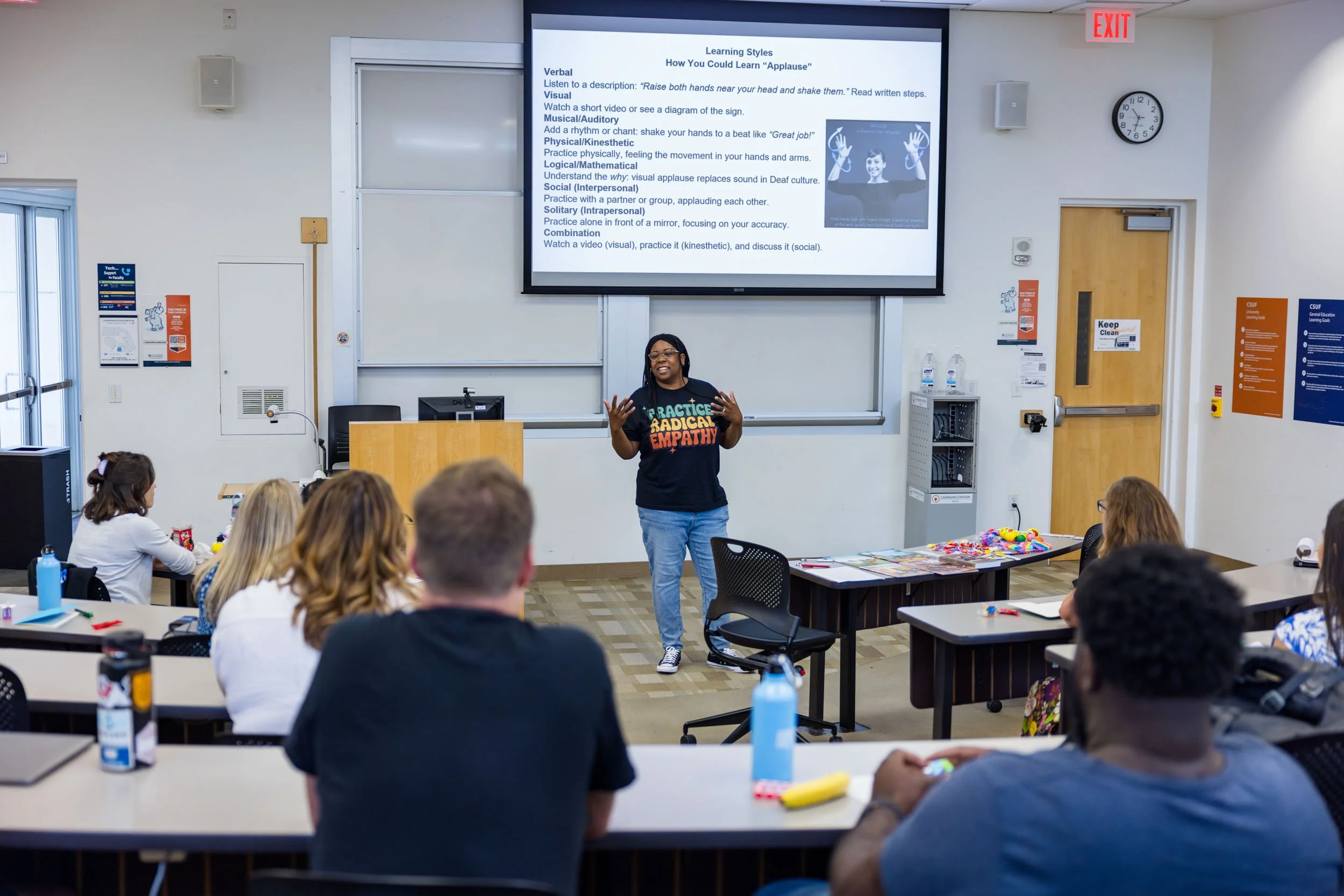A classroom with students sitting at desks, listening to a woman standing at the front. The woman is wearing a black T-shirt with colorful text and is giving a lecture about learning styles, as shown on the large screen behind her. The room has poste