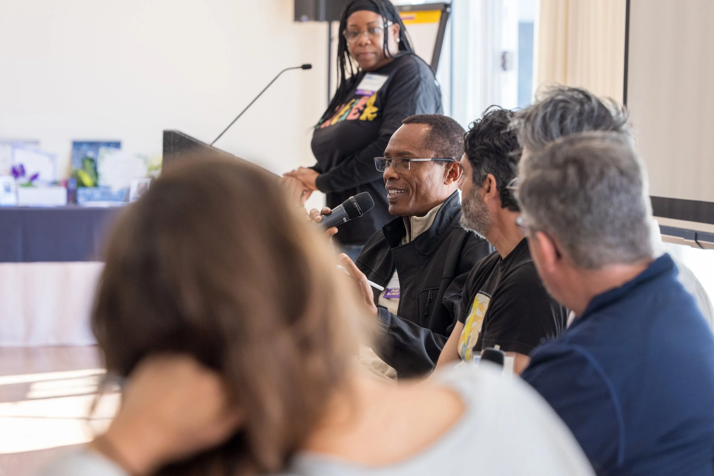 Man speaking into a microphone during a panel discussion, with other panelists and a woman standing behind him.