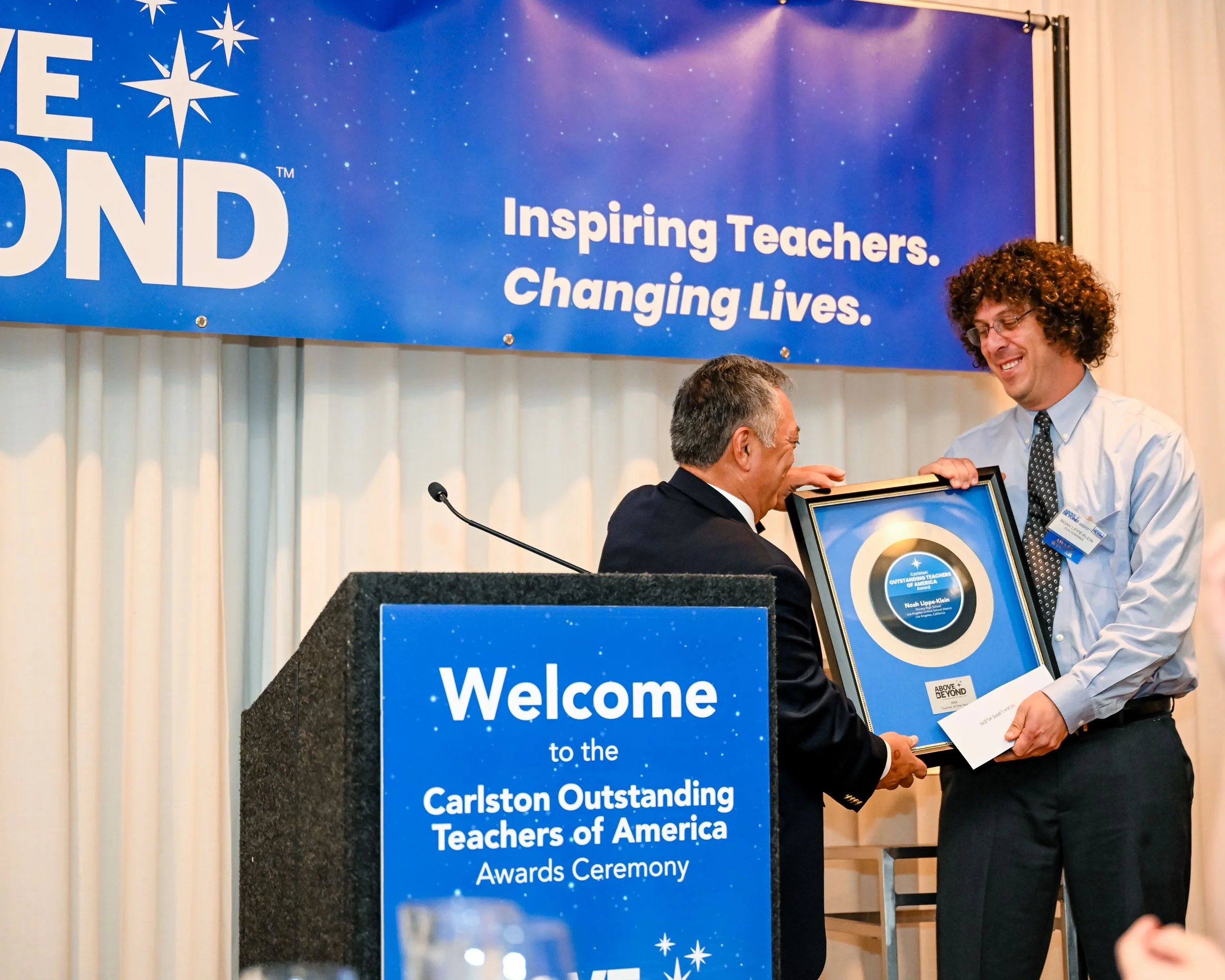 A man in a suit receiving an award from a man in a dress shirt at a ceremony with a blue banner in the background that reads 'Inspiring Teachers. Changing Lives.' and a sign in the foreground that says 'Welcome to the Carlston Outstanding Teachers of America Awards Ceremony.'