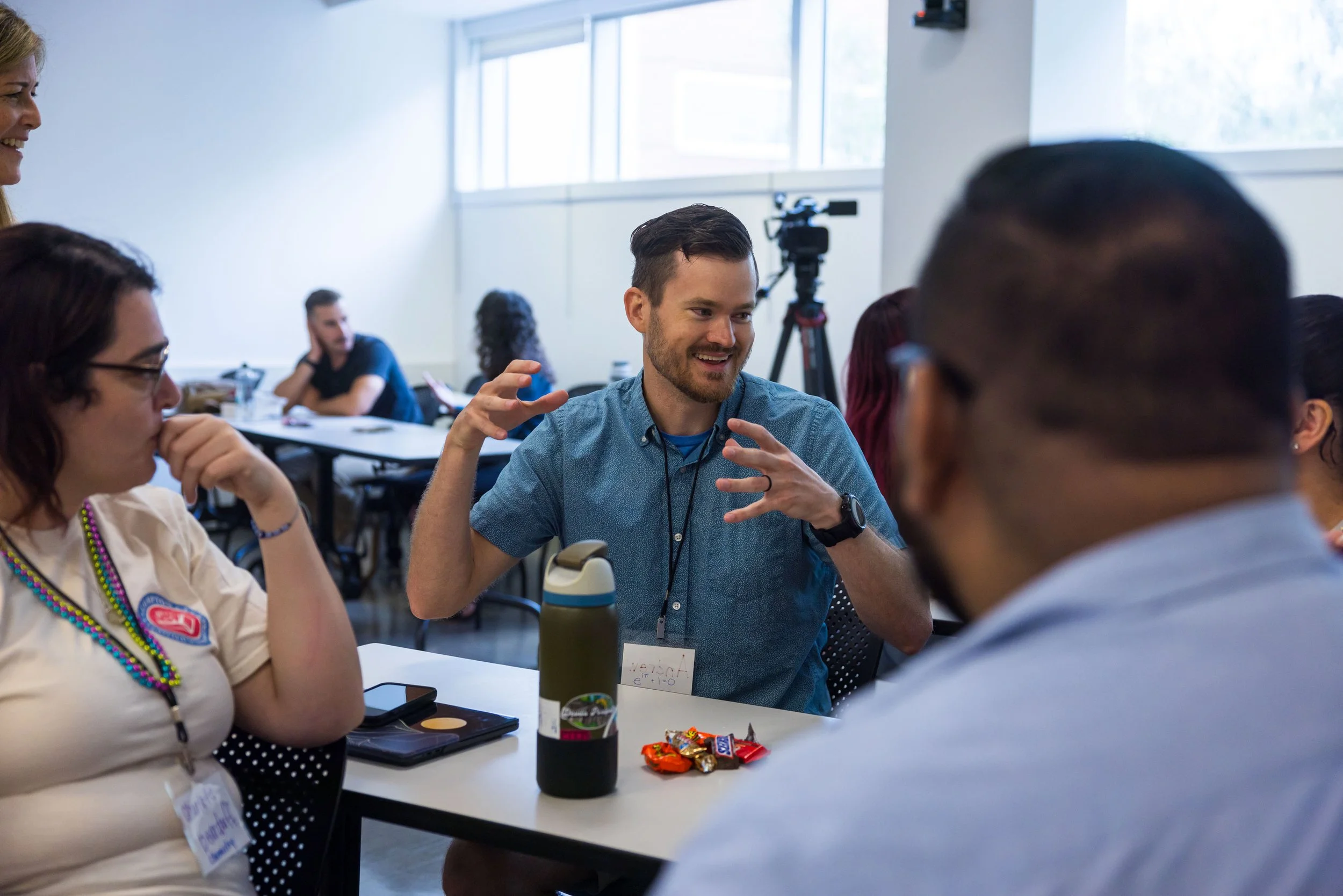 People sitting at a table having a conversation in a bright room. One man is smiling and gesturing while speaking, with snacks and a water bottle on the table.