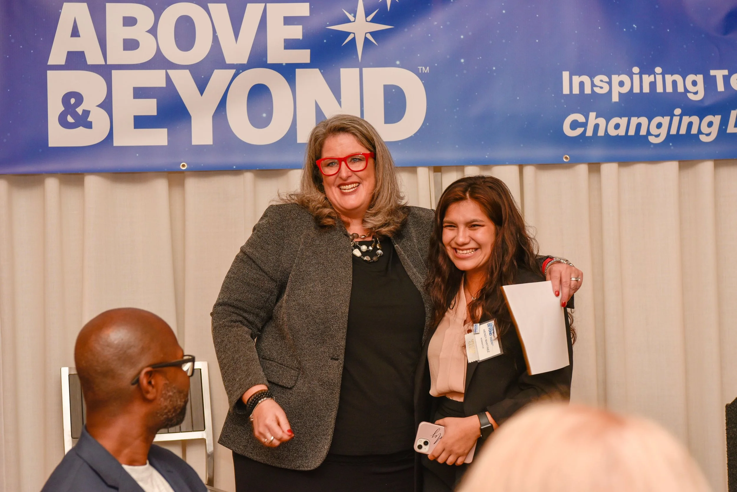 Two women smiling and posing for a photo at an event, with a blue banner reading 'Above & Beyond' behind them. One woman has gray hair, wears red glasses and a dark blazer, and the other has long dark hair, a black blazer, and is holding a book and a phone. A man with glasses and a bald head is sitting in the foreground.
