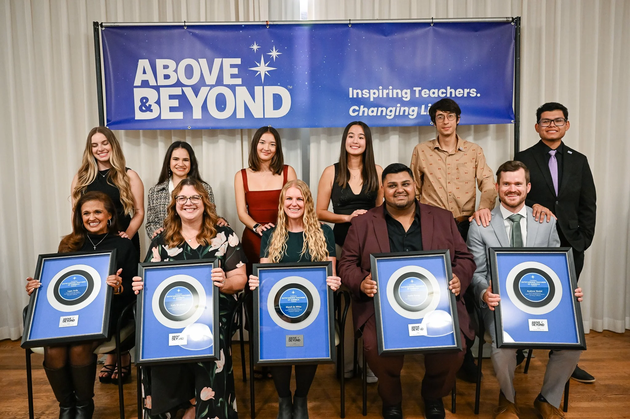 Group of ten diverse people posing with awards in front of a blue banner that reads 'Above & Beyond' and 'Inspiring Teachers, Changing Lives' in a room with white curtains.