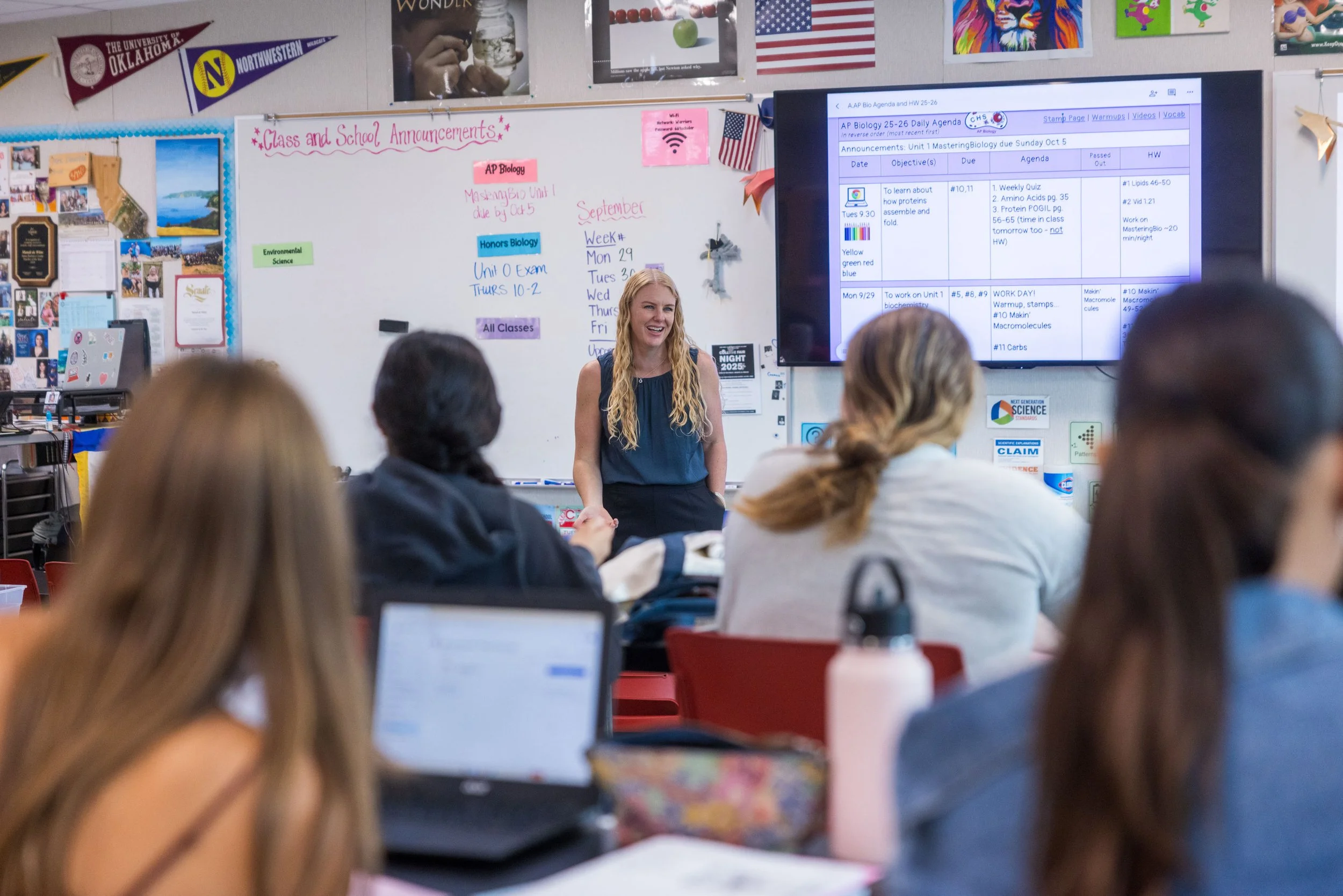 A classroom with students seated at desks facing a teacher. The teacher is smiling, standing near a whiteboard with colorful notes and a large screen displaying a lesson plan. The classroom is decorated with various posters and flags.