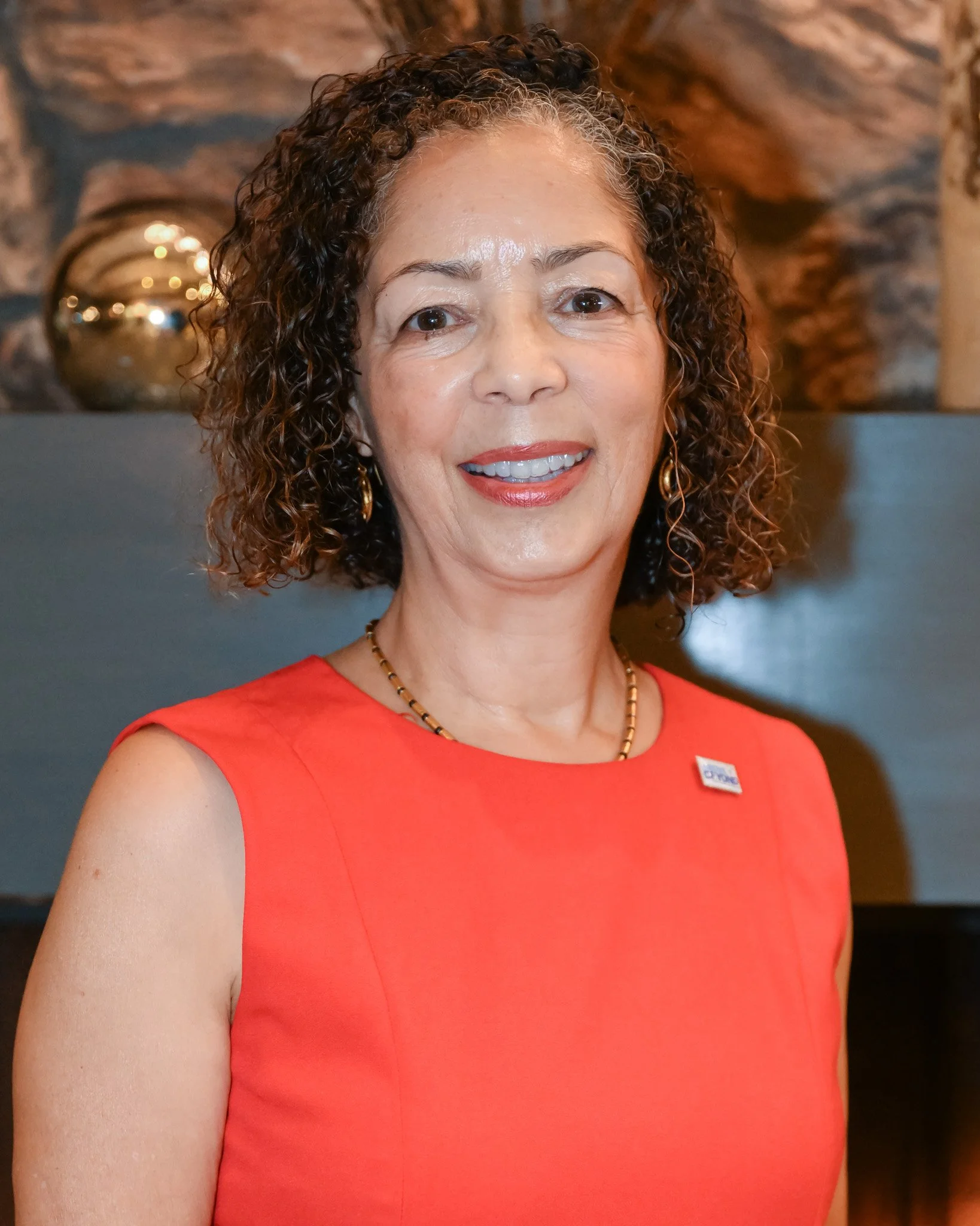 A woman with curly hair wearing a sleeveless red dress, gold earrings, and a necklace, smiling at the camera, with a decorative background.