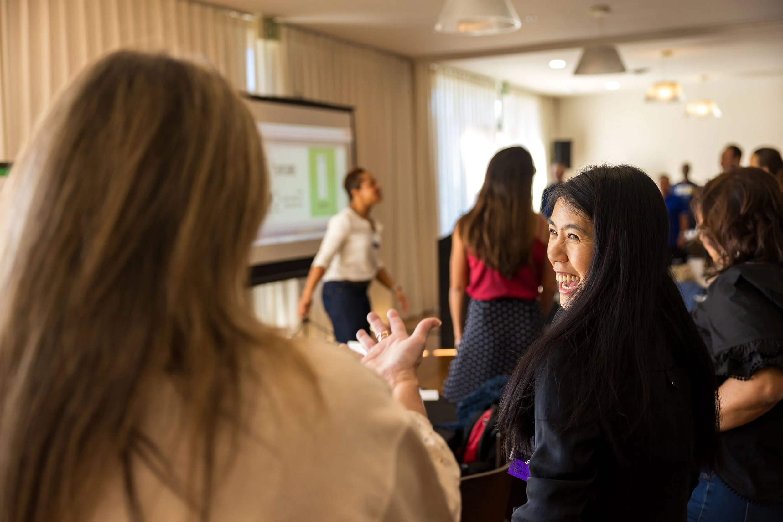 A woman with long black hair is smiling and talking to another person in a crowded conference room.