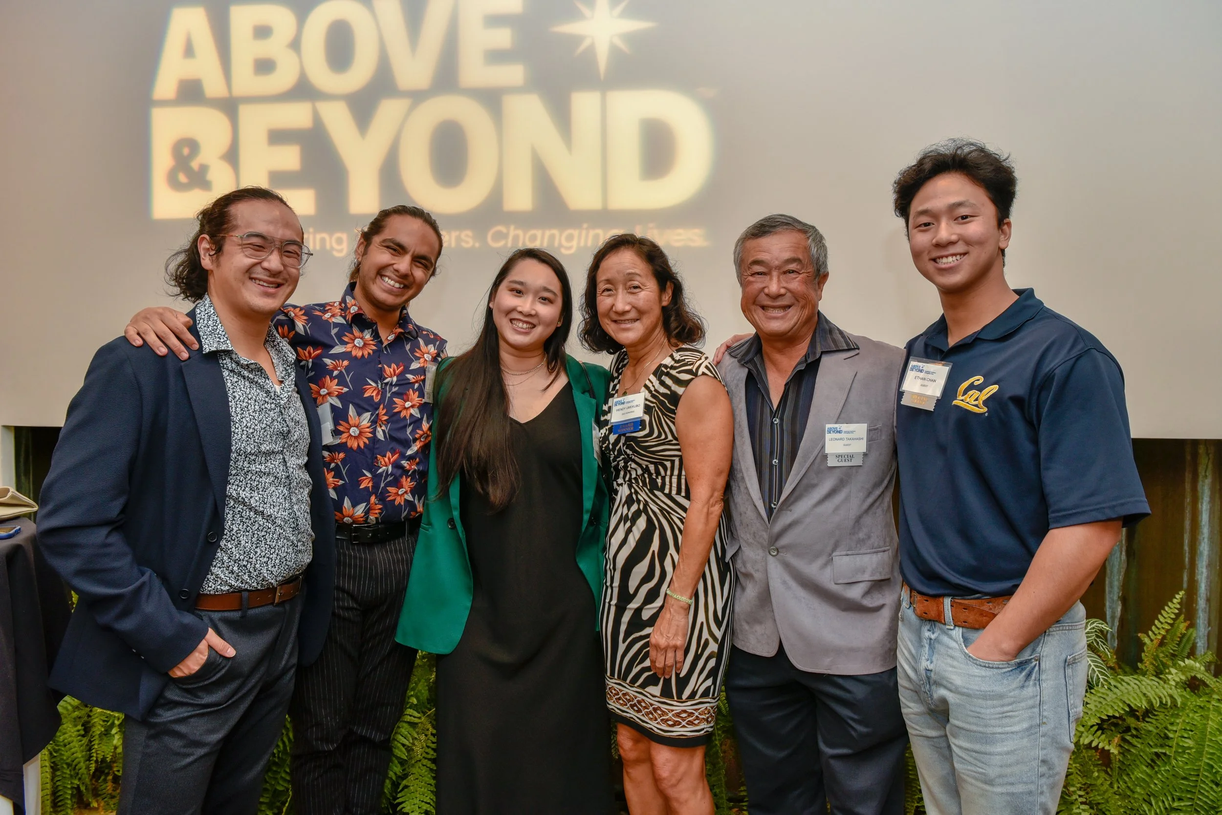 Group of seven diverse people smiling and posing together at an event, with a screen behind them displaying the words 'Above & Beyond' and a star symbol.