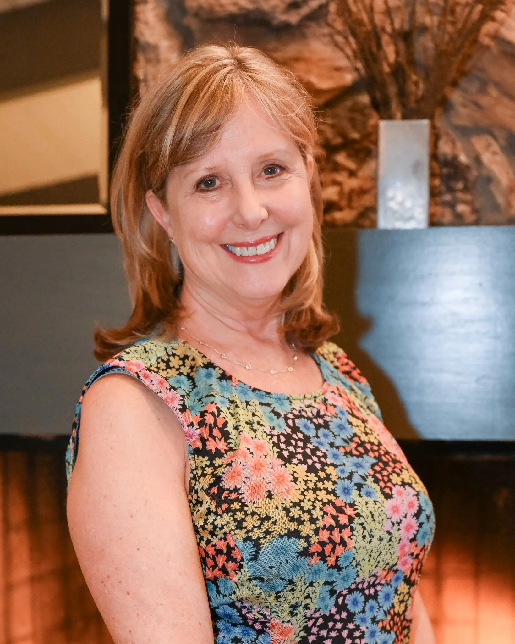 A smiling woman with shoulder-length red hair, wearing a sleeveless floral dress, standing indoors with a dark background featuring a piece of art and a vase.