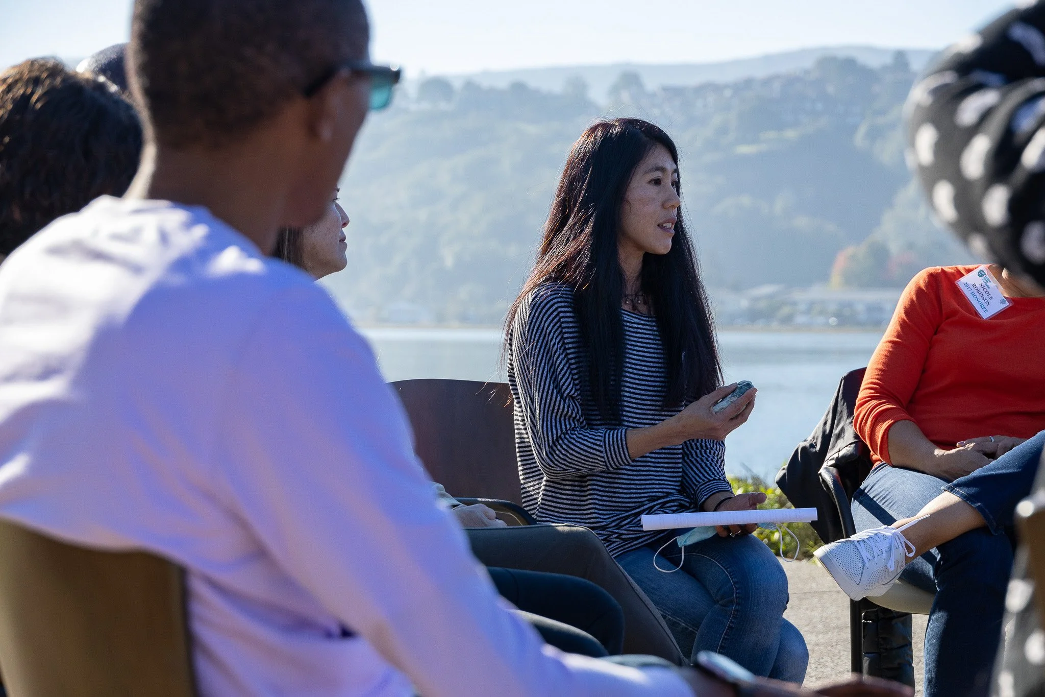 A group of people sitting outdoors near a body of water, engaged in a discussion or meeting, with a woman in a striped shirt holding a phone and a notebook.