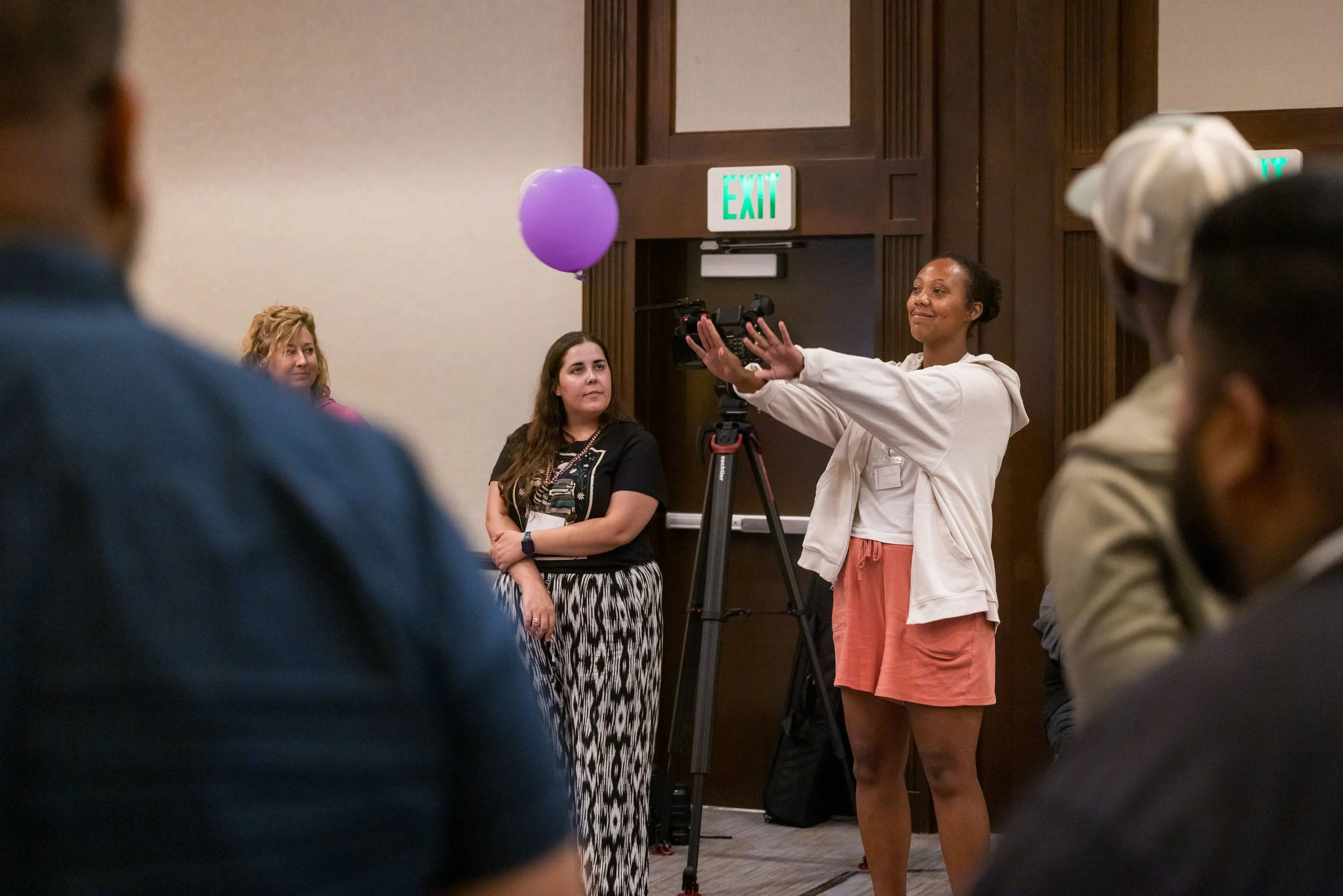 A woman standing in front of a camera with a purple balloon behind her, speaking to a group of people in a conference room with an exit sign above the door.