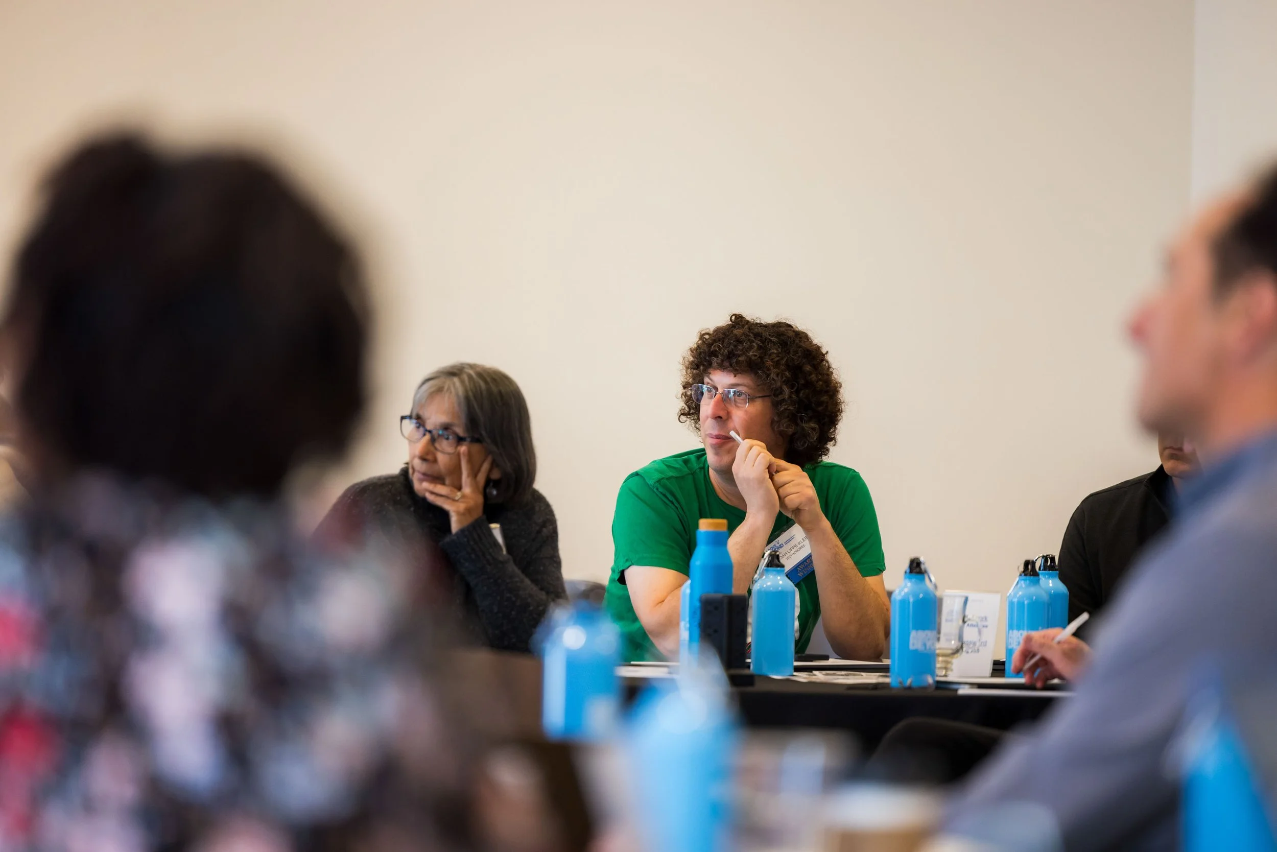 People sitting around a conference table. The woman on the left has gray hair and glasses, resting her chin on her hand. The man in the center has curly hair, glasses, and is wearing a green shirt. Bottles of water are on the table.