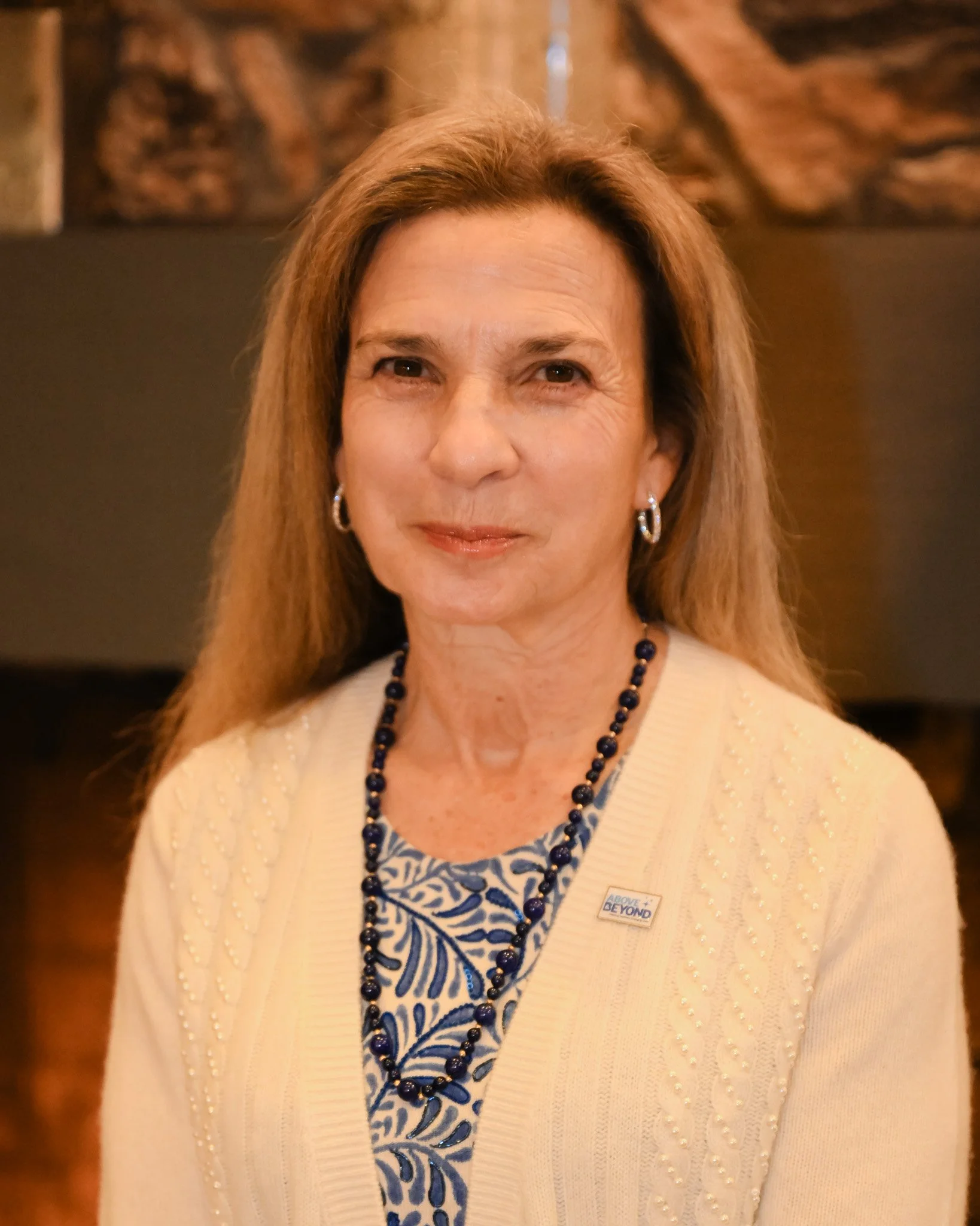 A middle-aged woman with long light brown hair, wearing a cream-colored cardigan with pearl embellishments, a blue and white patterned shirt, blue beaded necklace, and small hoop earrings. She is smiling slightly against a warm, blurred background.