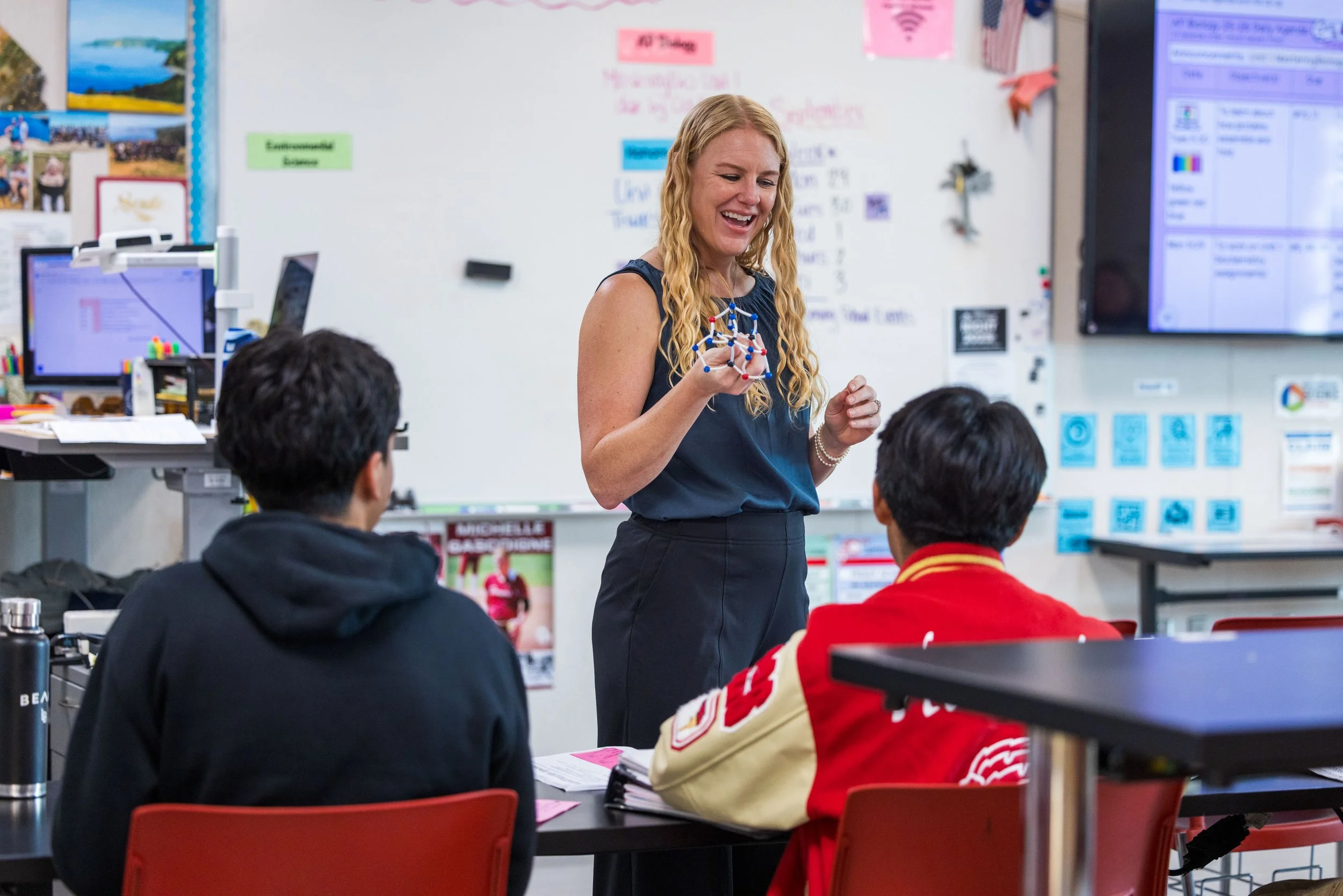 A woman teacher with blonde curly hair teaching two students in a classroom, holding a molecular model, with a whiteboard and computer screens in the background.