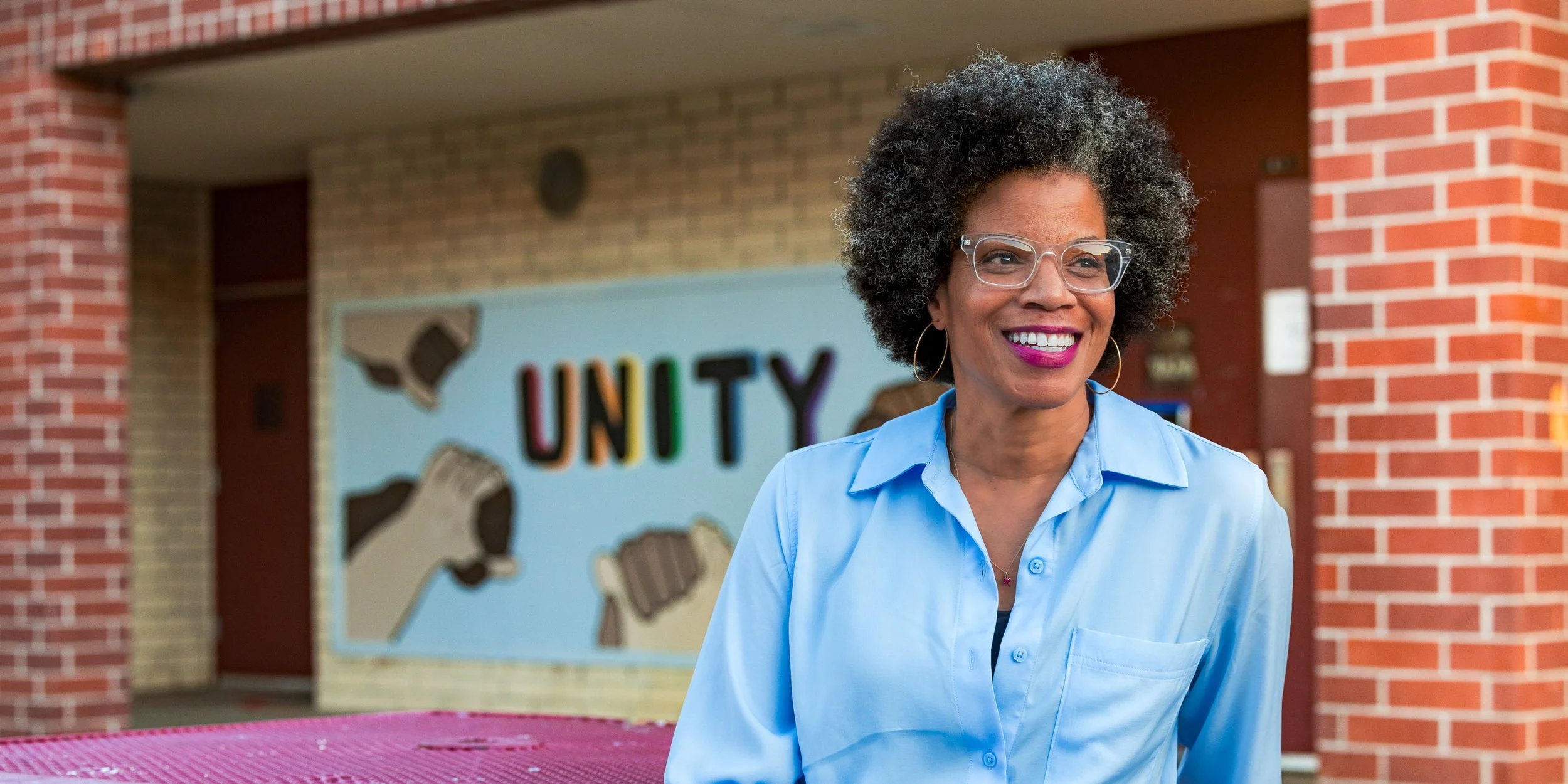 A woman with glasses and curly hair smiling outdoors in front of a mural reading "UNITY" and depicting hands coming together.