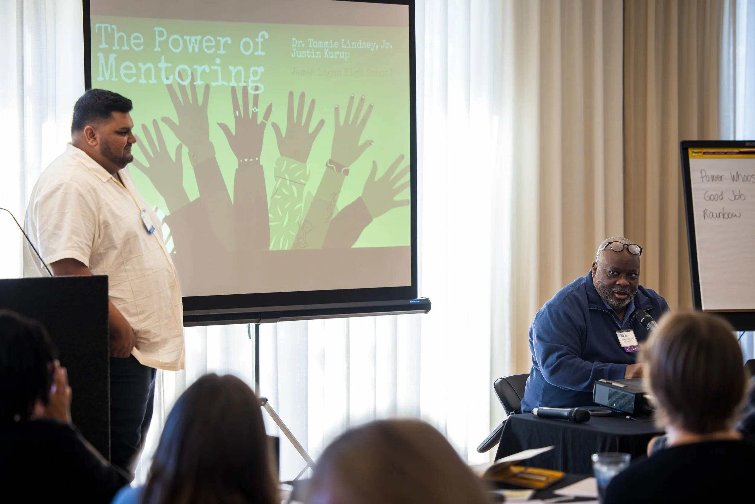 A man in a white shirt standing next to a presentation screen titled "The Power of Mentoring" with an illustration of raised hands. An older man in a blue jacket is sitting next to a microphone, speaking to an audience in a conference room.