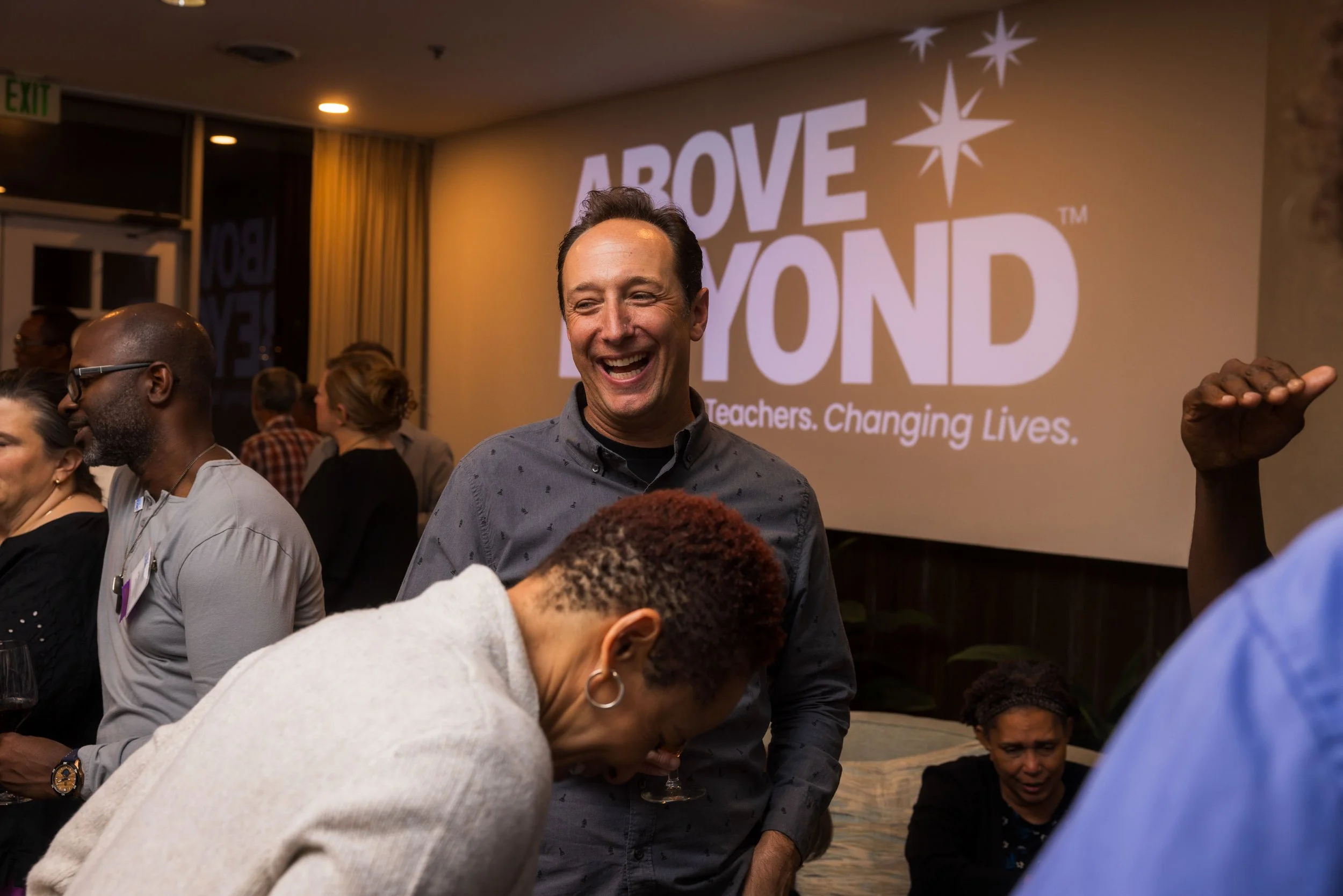 A group of people at an event with a large screen in the background displaying the words 'ABOVE BEYOND' and the tagline 'Teachers. Changing Lives.' One man in the center is smiling and laughing, wearing a gray shirt, while others are engaged in conversation.