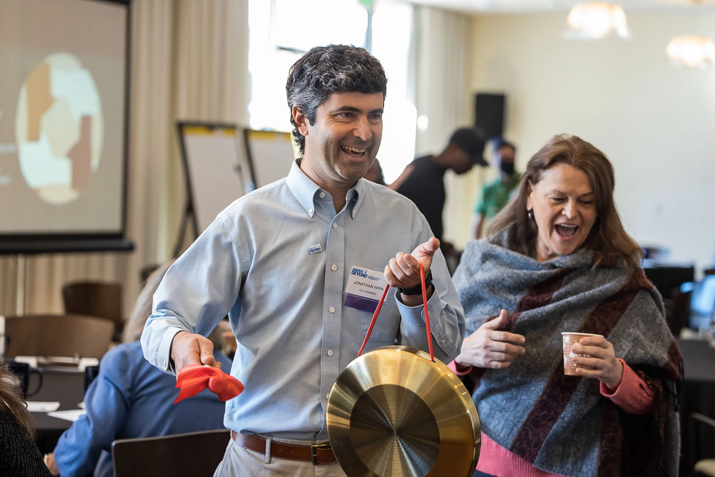 Man smiling and holding a gong at a conference or meeting, with woman beside him laughing and holding a cup, in a brightly lit room with others in the background.