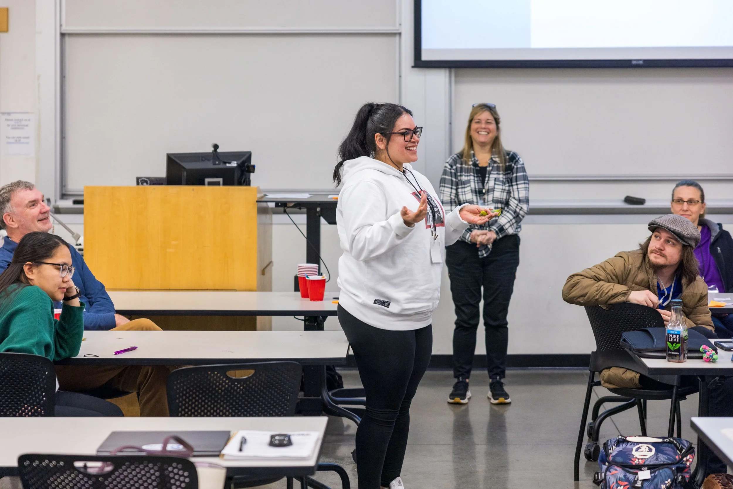 A woman in a white hoodie standing and speaking in front of an audience in a classroom, with four people seated and listening, two women standing behind her, and classroom equipment visible.