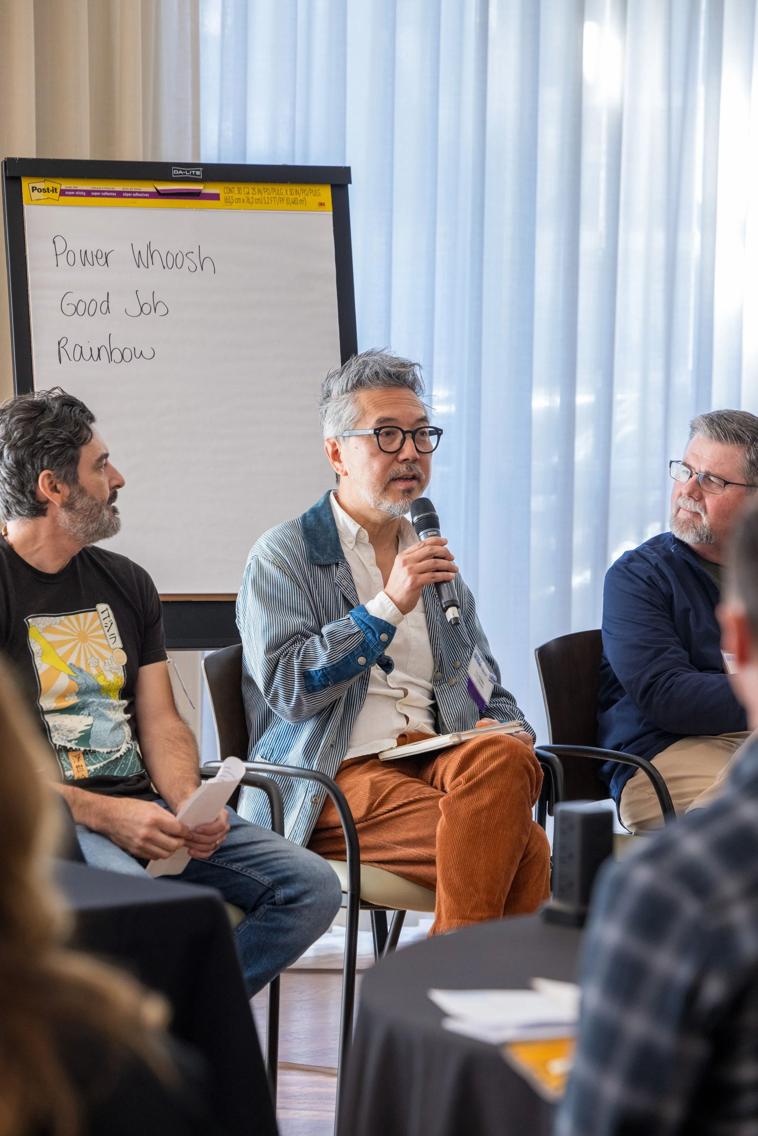 A man with glasses holding a microphone speaking to a group at a panel discussion, with two other men seated beside him and a whiteboard in the background.