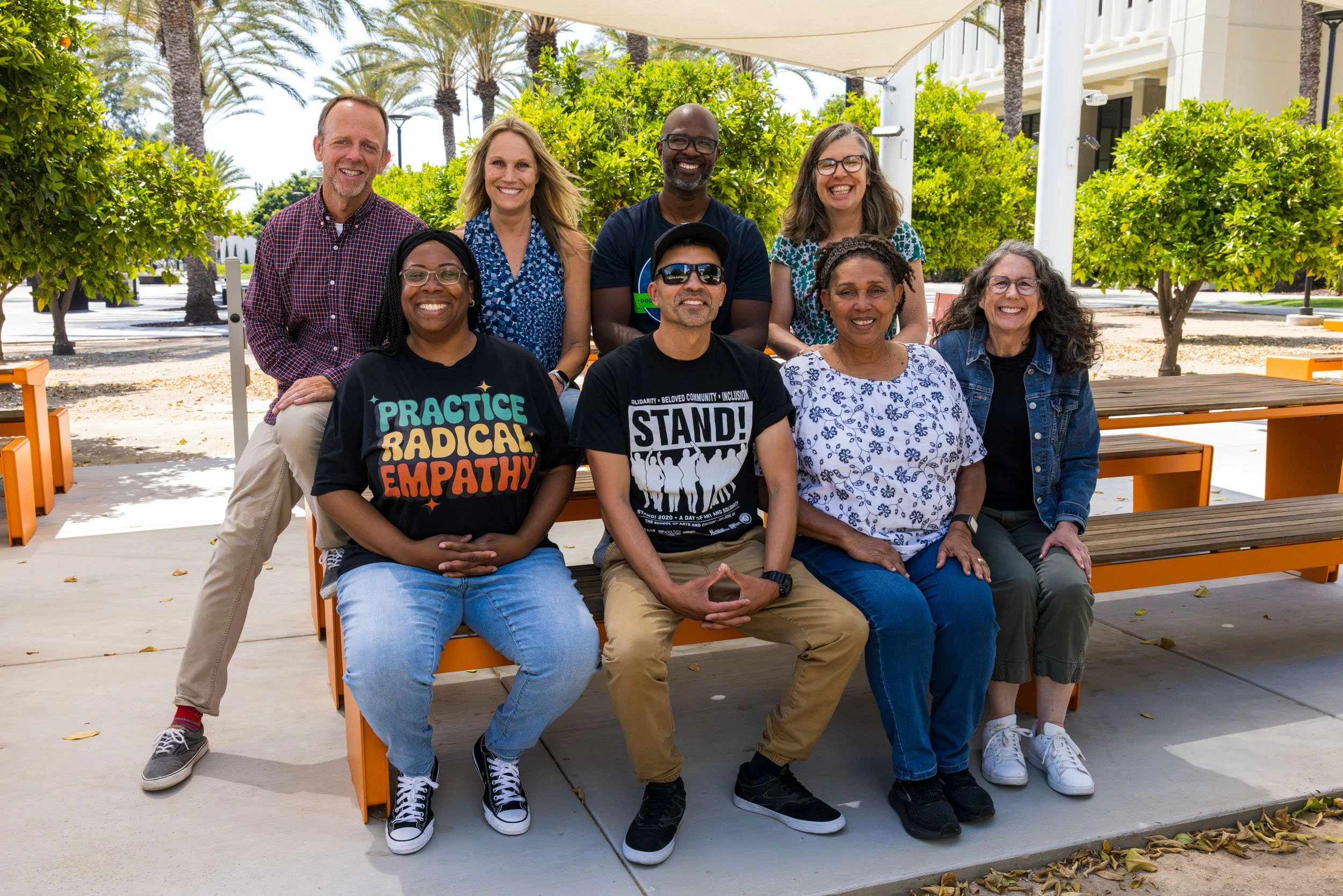 A group of nine diverse people smiling and posing for a photo outdoors on a sunny day, sitting and standing on orange benches with trees and palm trees in the background.